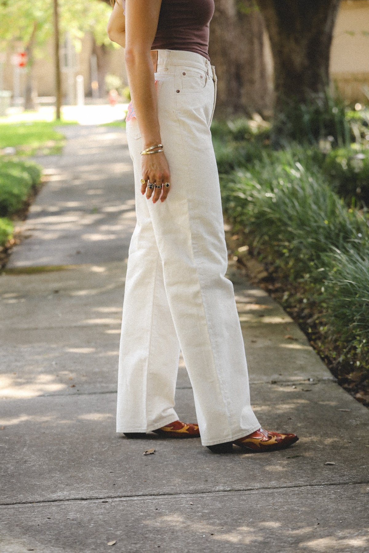 A person standing on a sidewalk outdoors wearing white wide-leg jeans, a dark brown sleeveless top, and brown patterned loafers, with trees and greenery in the background.