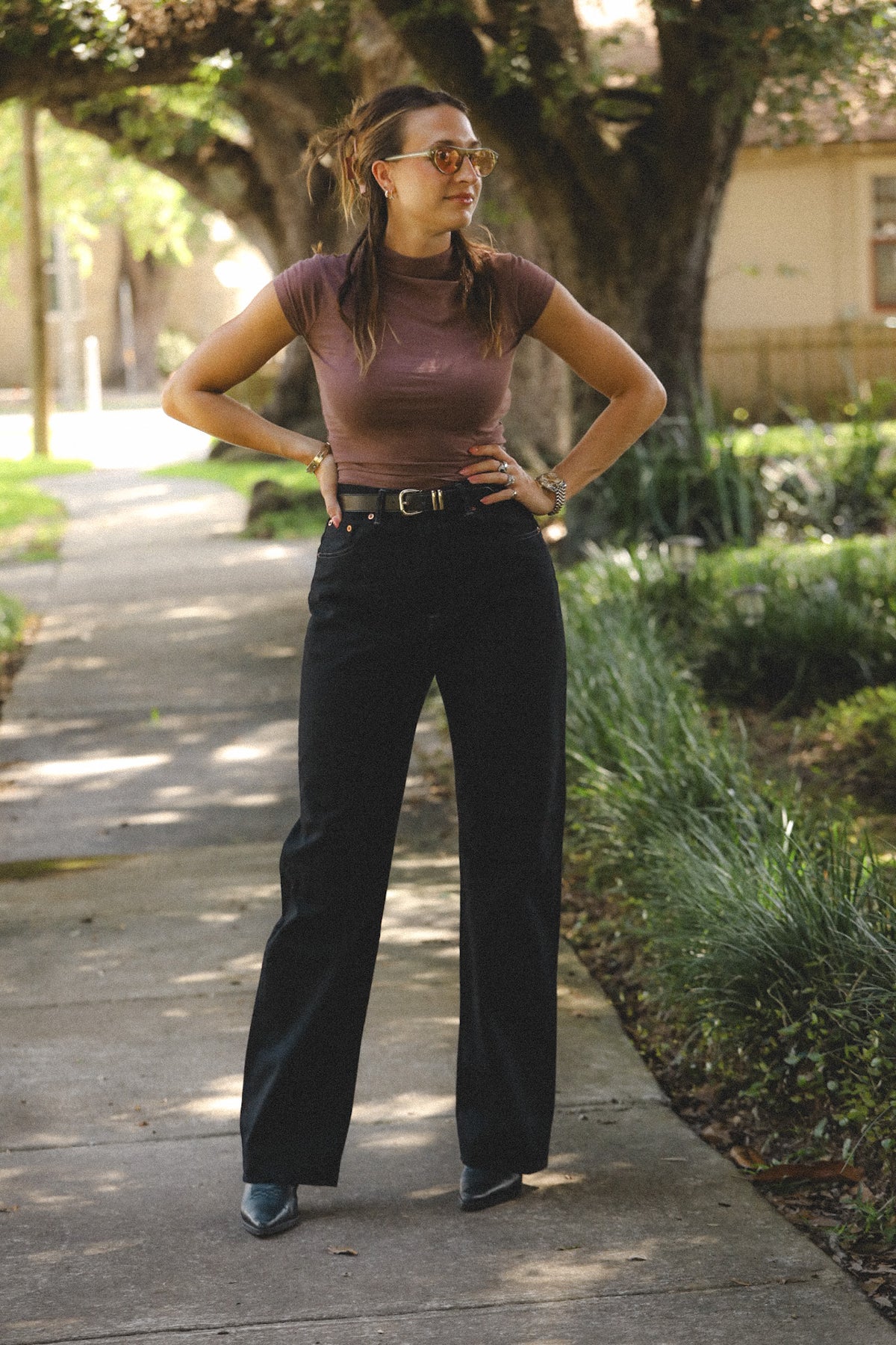 A woman stands on an outdoor sidewalk wearing a fitted short-sleeve mauve-brown mock-neck top, black wide-leg pants, a black belt, and dark pointed shoes.