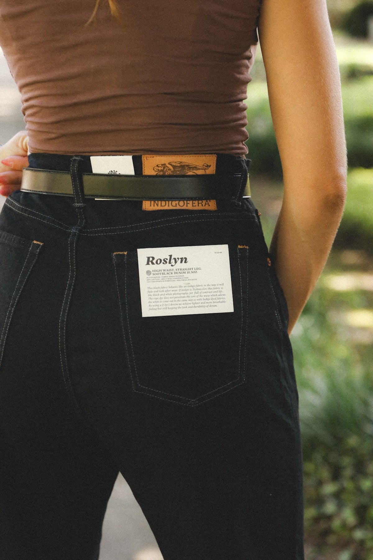A person wearing black denim jeans with a brown belt and a white paper label in the back pocket, photographed outdoors with a blurred green background.