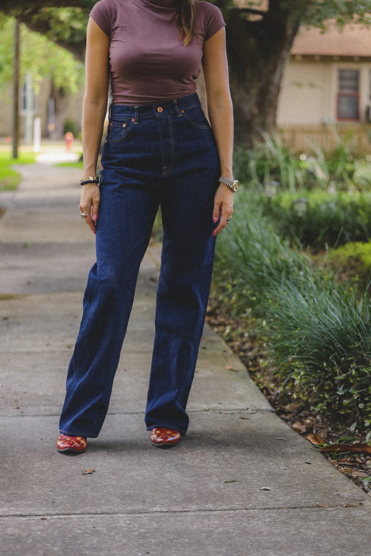 A person standing on a sidewalk wearing dark blue high-waisted wide-leg jeans, a mauve turtleneck top, and red-and-white patterned shoes, with trees and greenery in the background.
