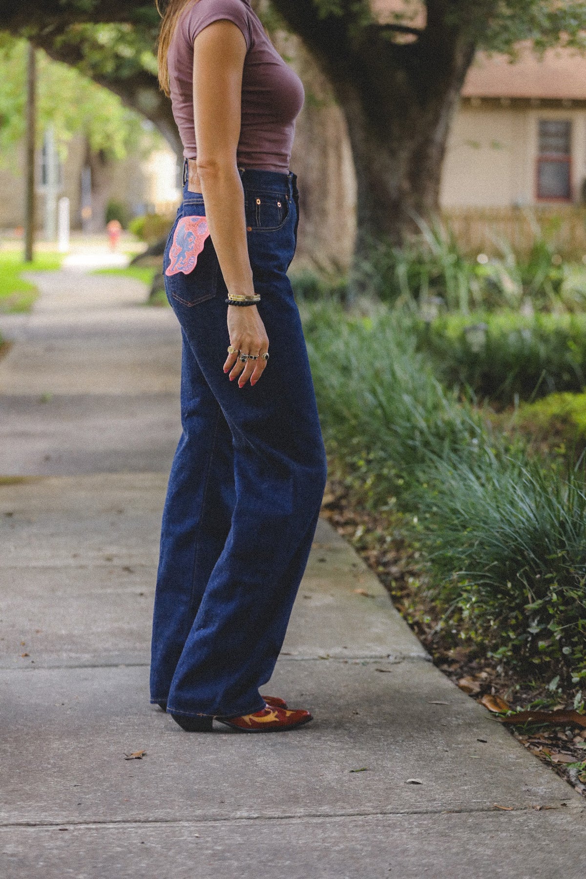 A person standing on a sidewalk outdoors, wearing dark blue wide-leg jeans with a pink and white patterned patch on the back pocket, a mauve short-sleeve top, and brown shoes.