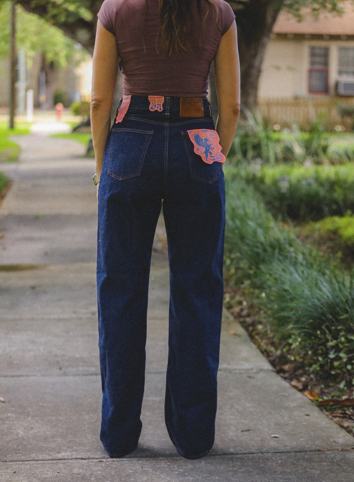 A person viewed from behind wearing dark blue wide-leg jeans with colorful embroidered patches on the back pockets, standing on a sidewalk outdoors.