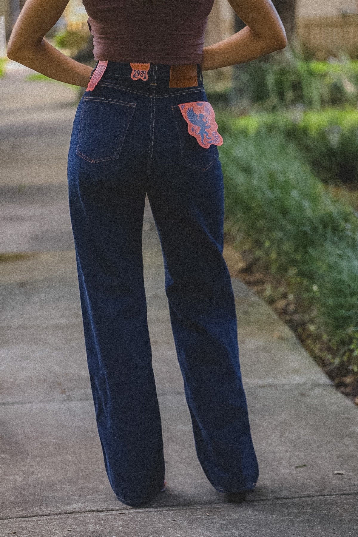 A person viewed from behind wearing dark blue wide-leg jeans with pink decorative patches on the back pockets, standing on a concrete sidewalk outdoors.