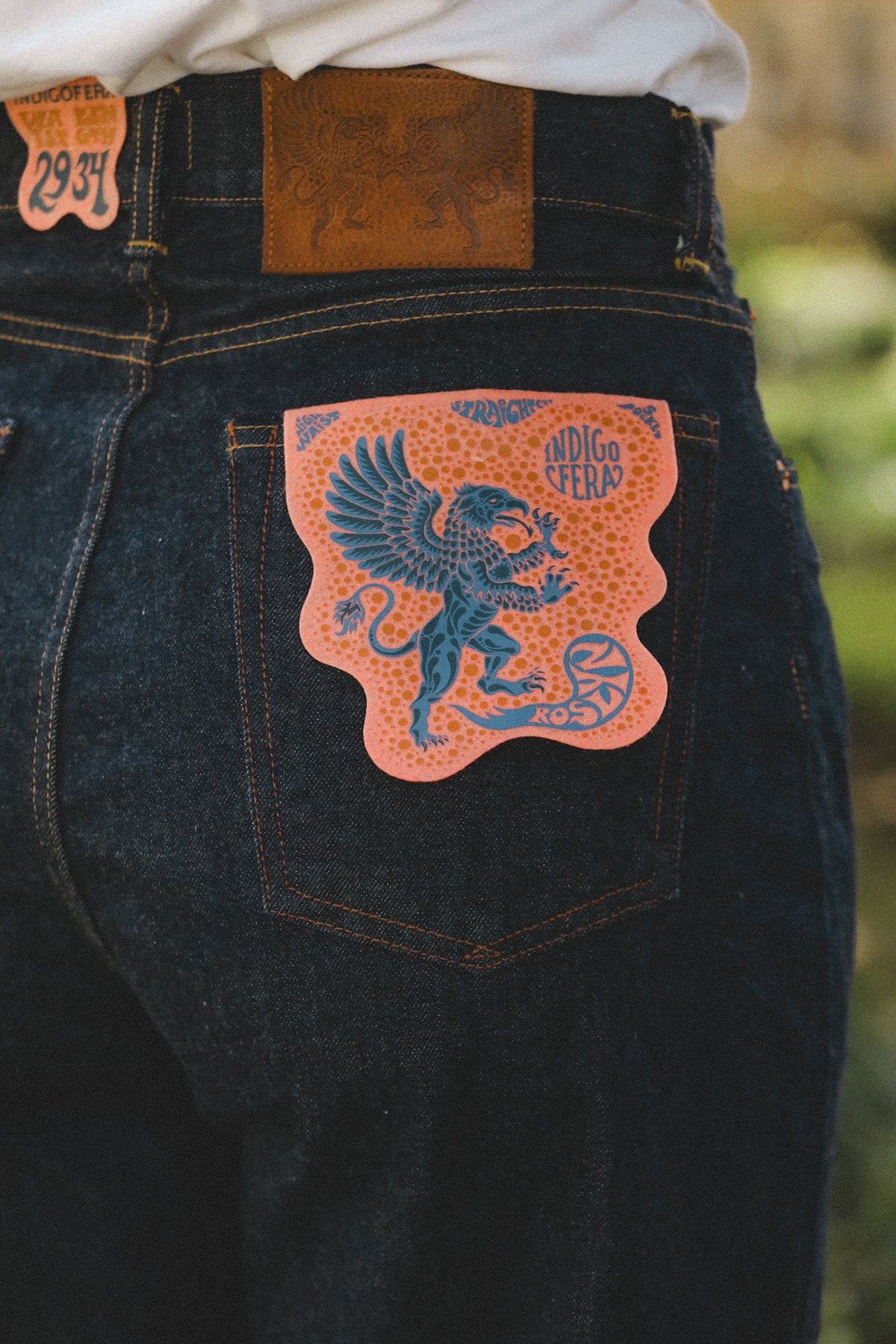 Close-up of the back waistband of dark indigo denim jeans, showing a tan leather patch, a stitched leather patch reading '23TH', and a prominent orange back pocket patch featuring a blue griffin and the text 'INDIGO STEEL'.