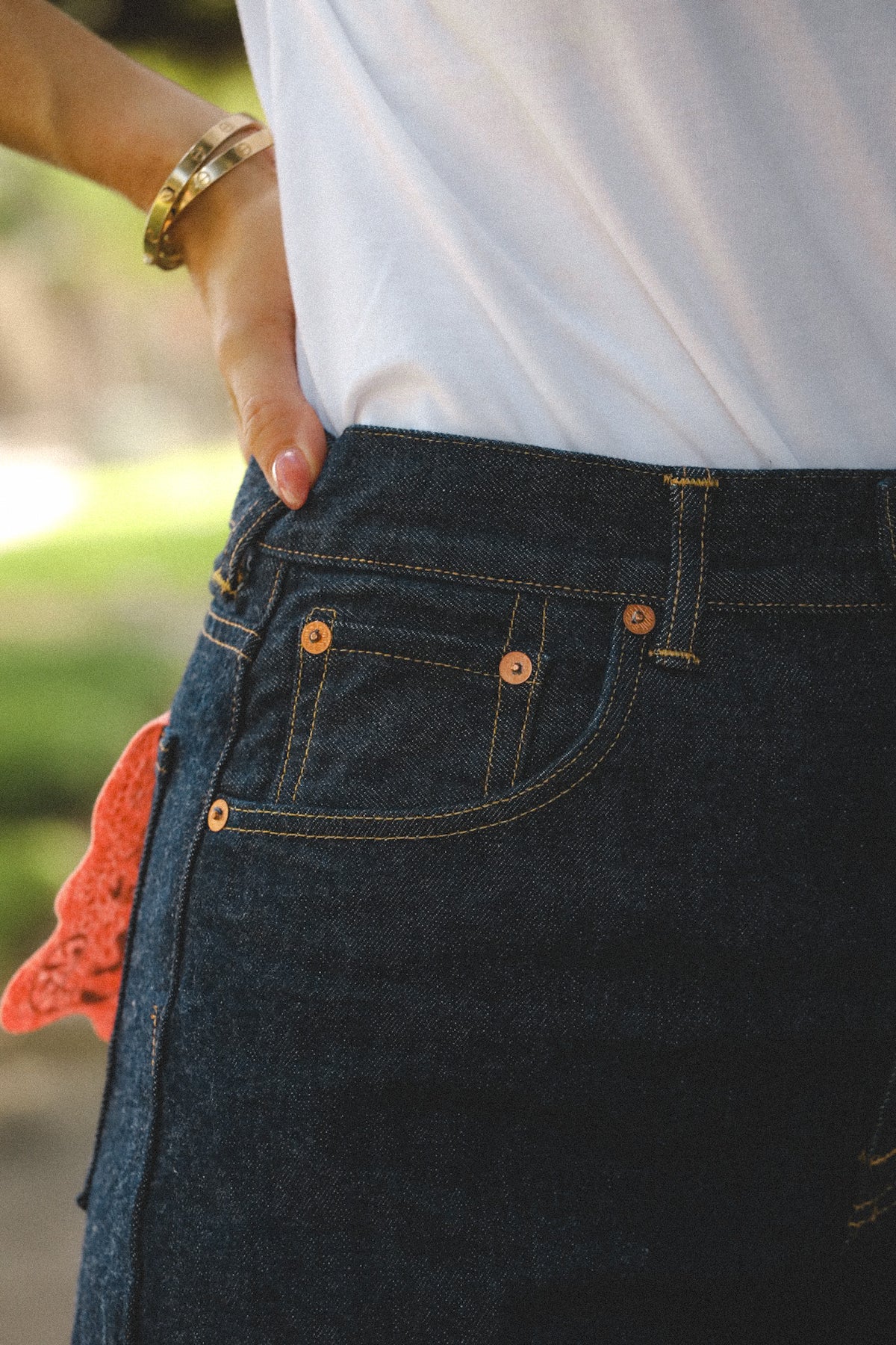 Close-up of a person wearing dark indigo denim jeans with a white top, showing the back pocket, copper-toned rivets, and a gold bracelet on the wrist.