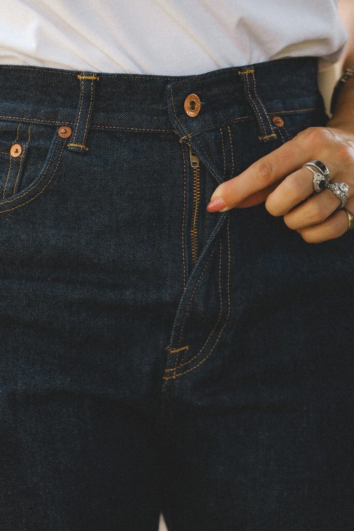 Close-up of a person wearing dark indigo denim jeans, pointing at the front zipper with a silver ring on their finger.