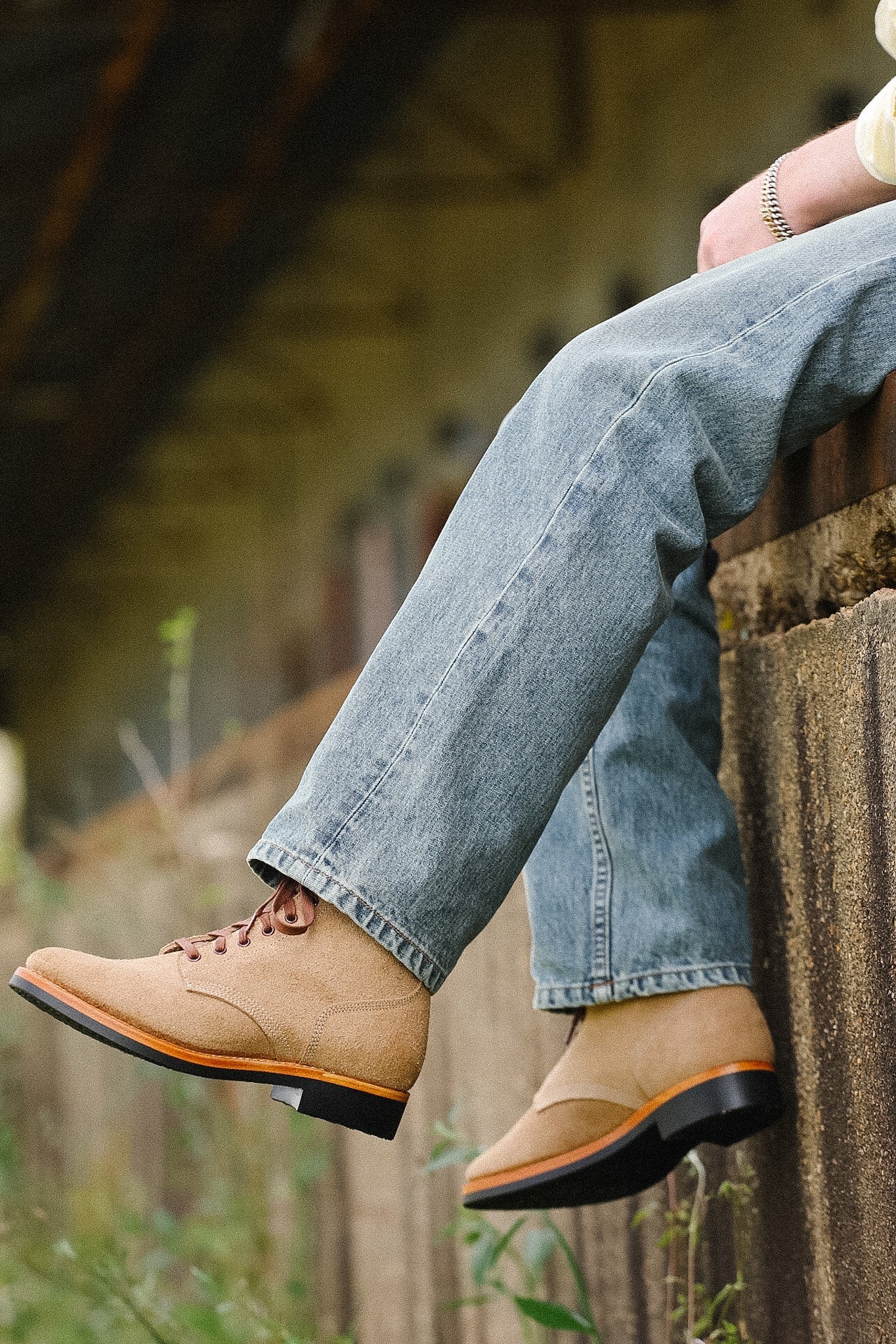Person wearing brown leather shoes with orange soles and blue jeans, sitting on a wooden bench outdoors.