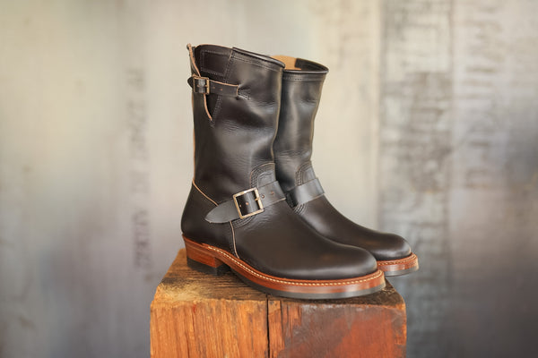Pair of black leather boots with buckles on a wooden block against a textured gray background
