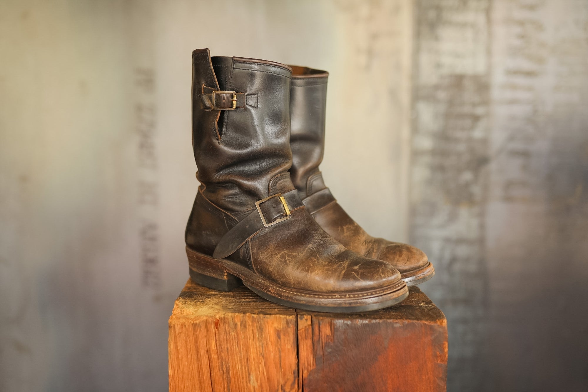 Pair of brown leather boots with buckles on a wooden block against a textured wall.