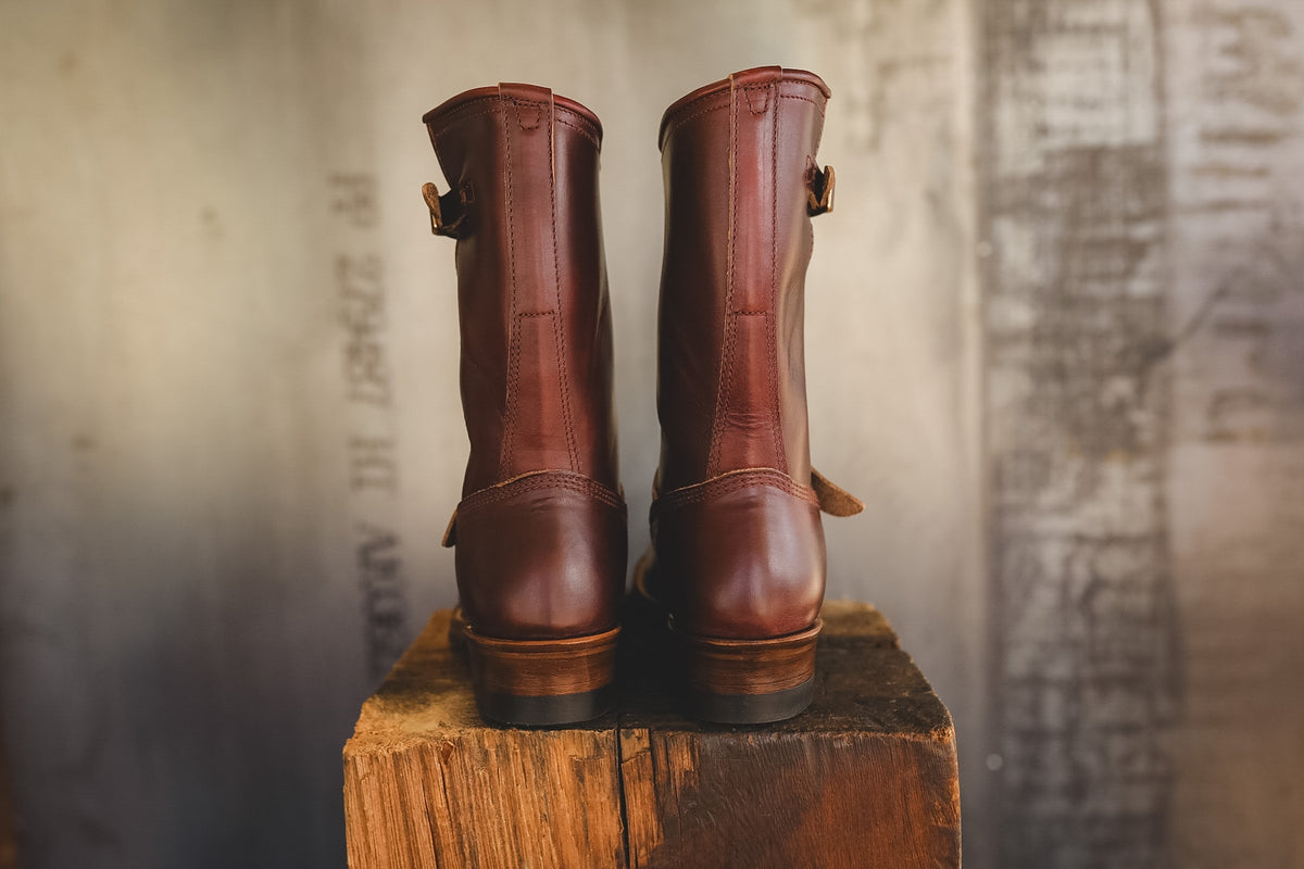 A pair of dark brown leather mid-calf boots with side buckle details, standing side-by-side on a rustic wooden block against a textured, warm-toned background.