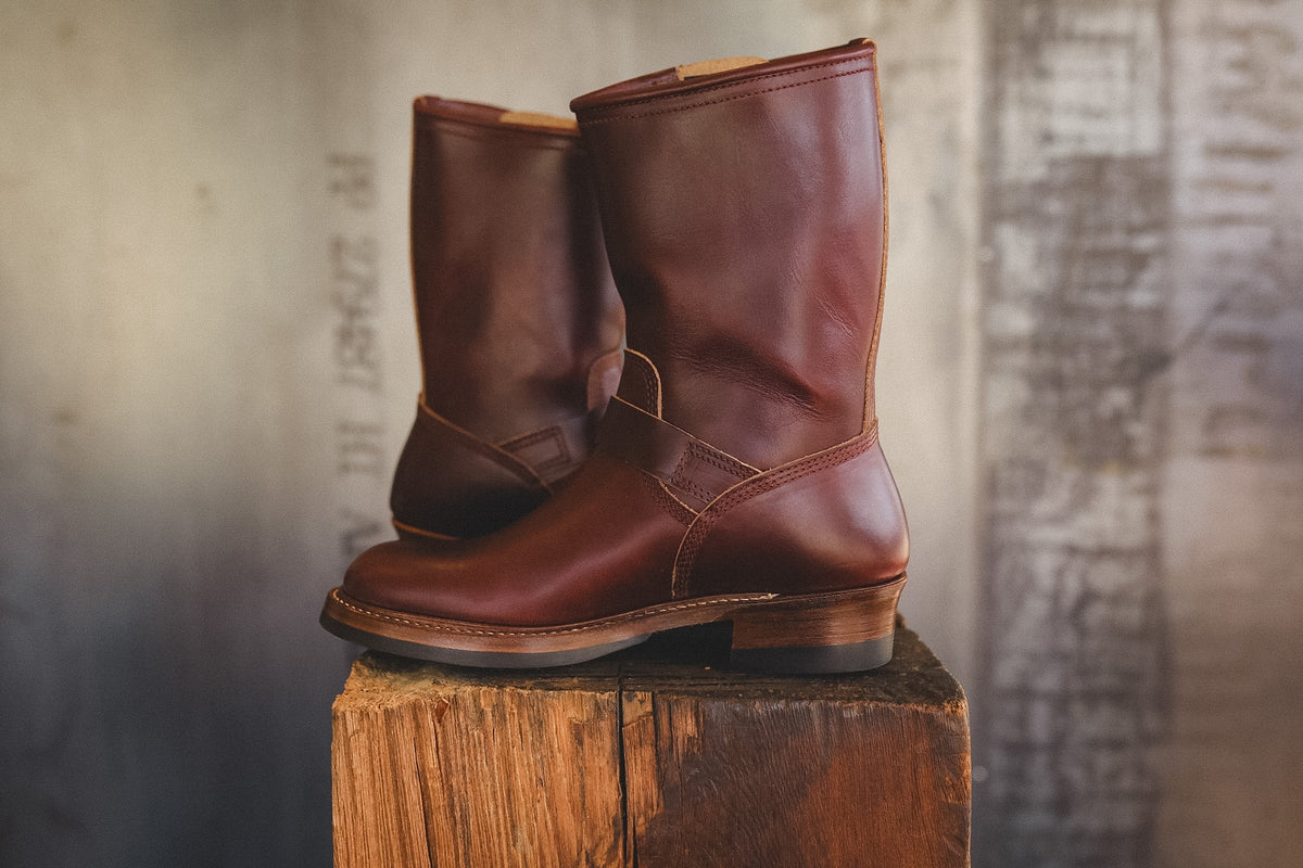 A pair of mid-calf brown leather boots with a lace-up front, displayed on a rustic wooden block against a textured, blurred gray background.