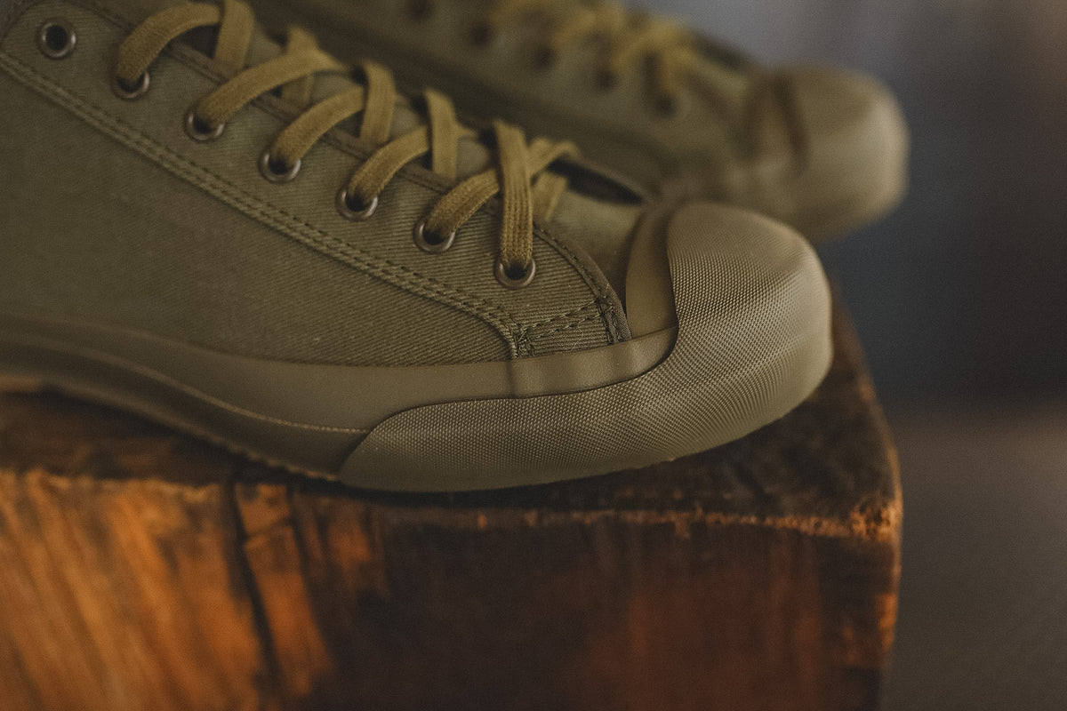 Up close image of a olive green sneaker on a wood block with a grey background 
