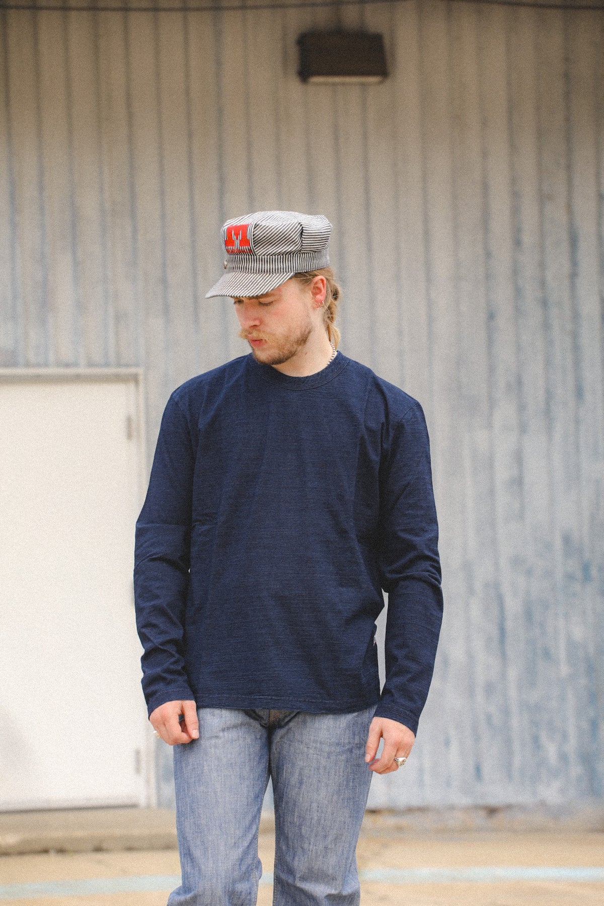 A man wearing a navy blue long-sleeve crewneck shirt, blue jeans, and a gray cap with a red logo, standing in front of a corrugated metal building.