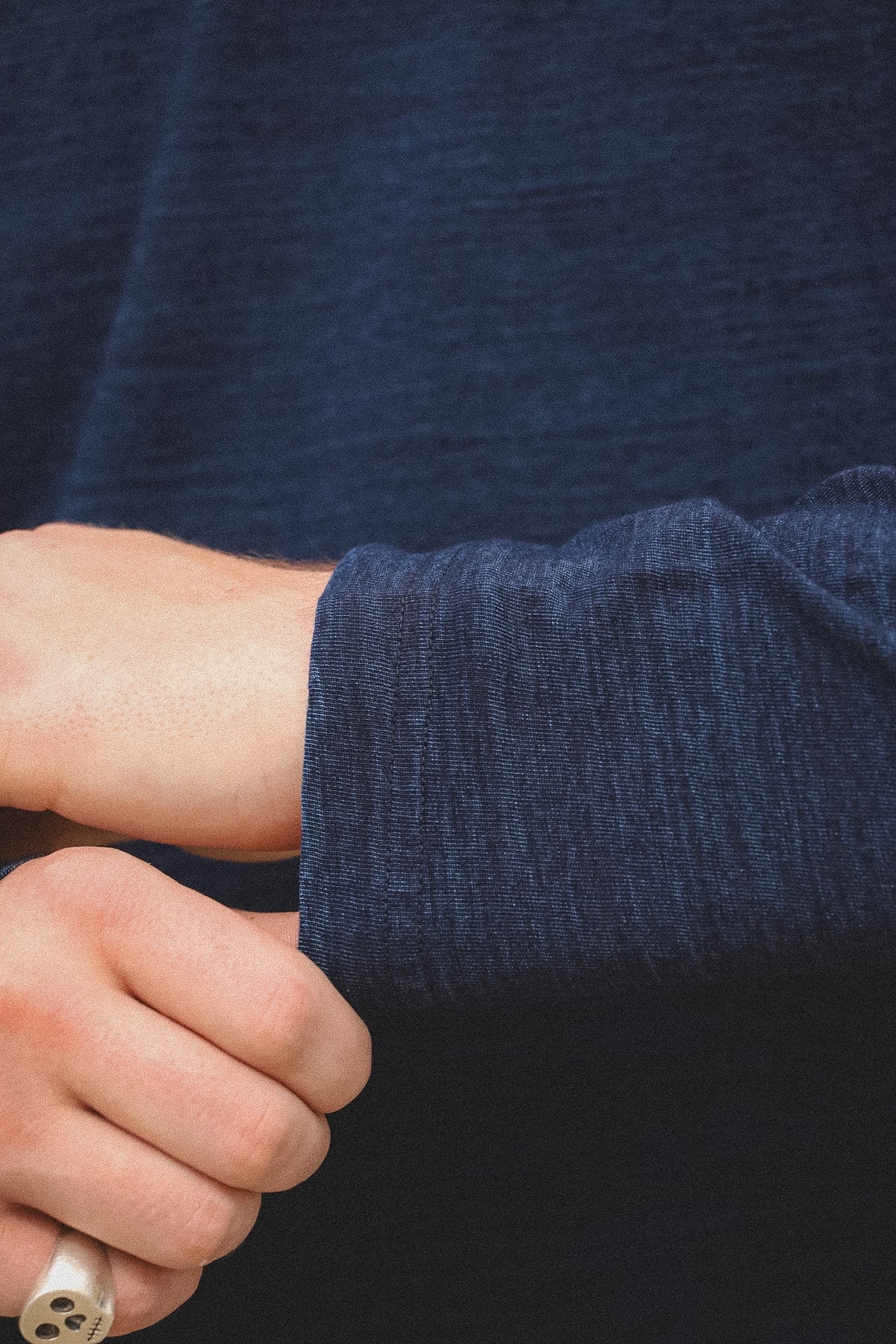 Close-up of a person's hands rolling up the sleeve of a dark navy blue textured knit sweater, with a cigarette holder in one hand.