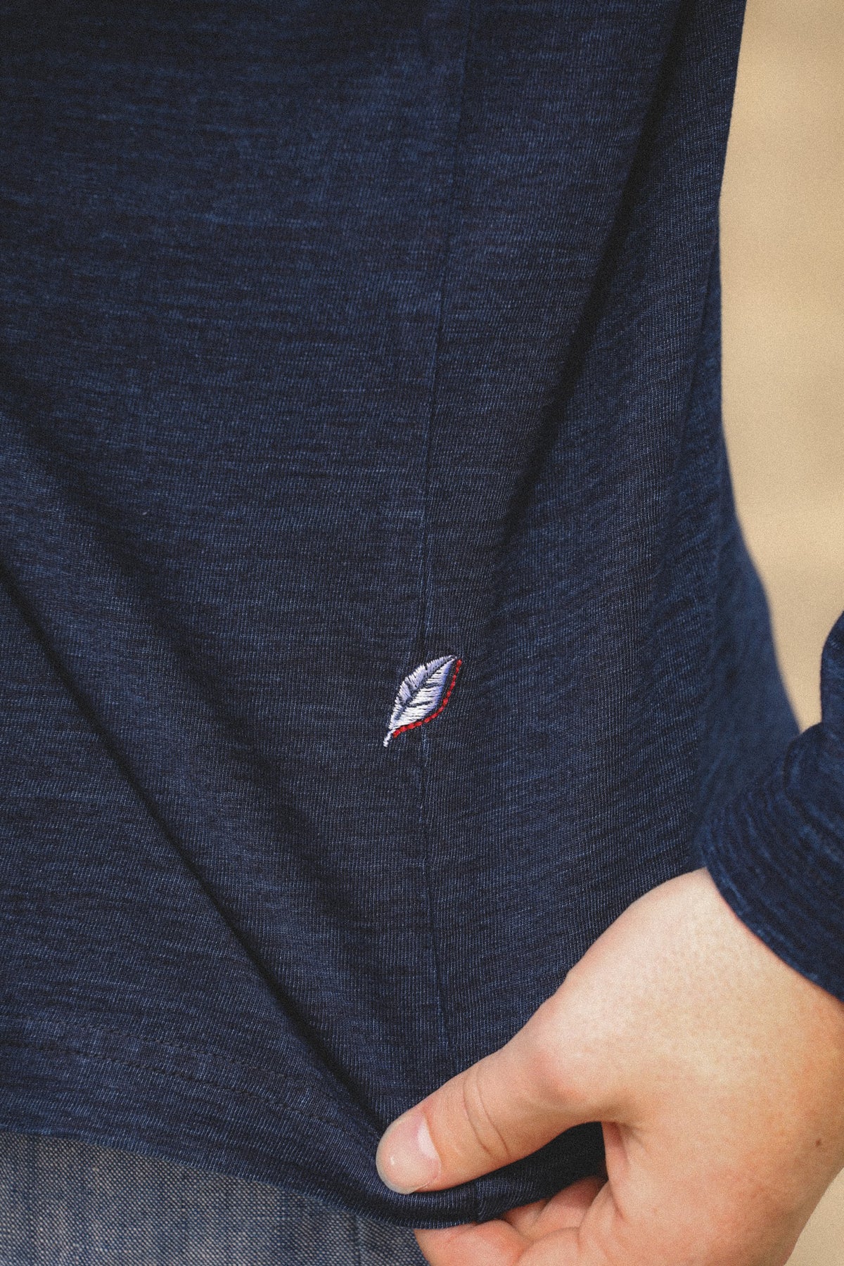 Close-up of a person wearing a dark navy heathered long-sleeve shirt, pulling the hem slightly to show a small embroidered feather logo in red, white, and blue.
