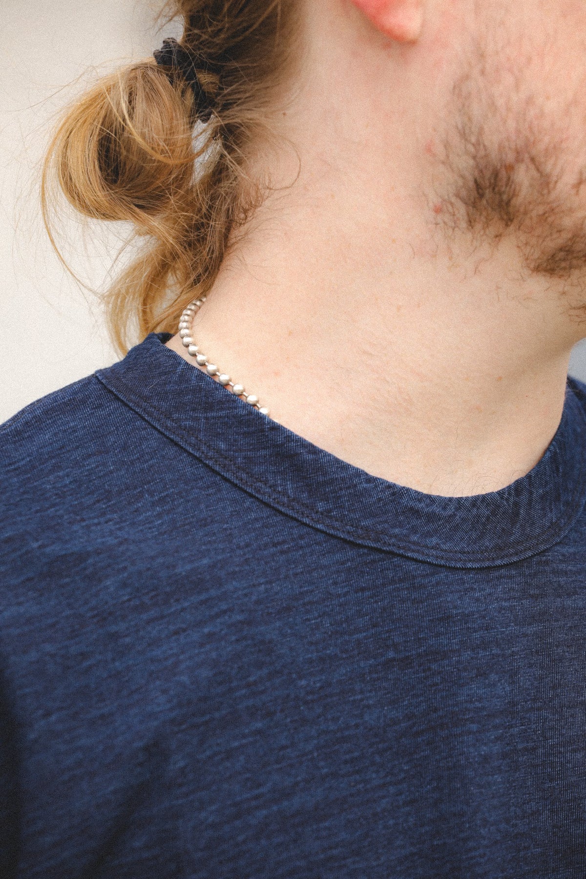 A close-up lifestyle shot of a person wearing a delicate silver beaded necklace and a dark blue heathered t-shirt.
