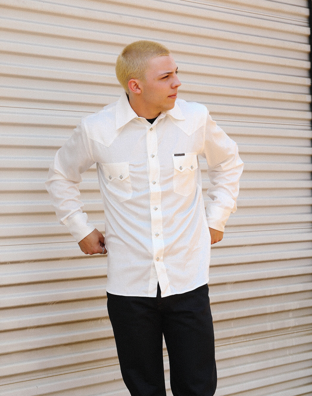 Man wearing a white Lindell Tencel cotton long-sleeve shirt with black pants against a beige corrugated metal background