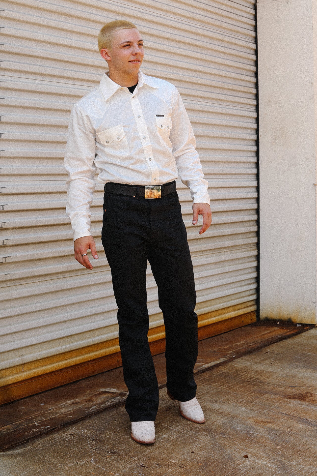Man wearing white Tencel cotton shirt, black jeans, white boots, and decorative belt standing by metal roll-up door