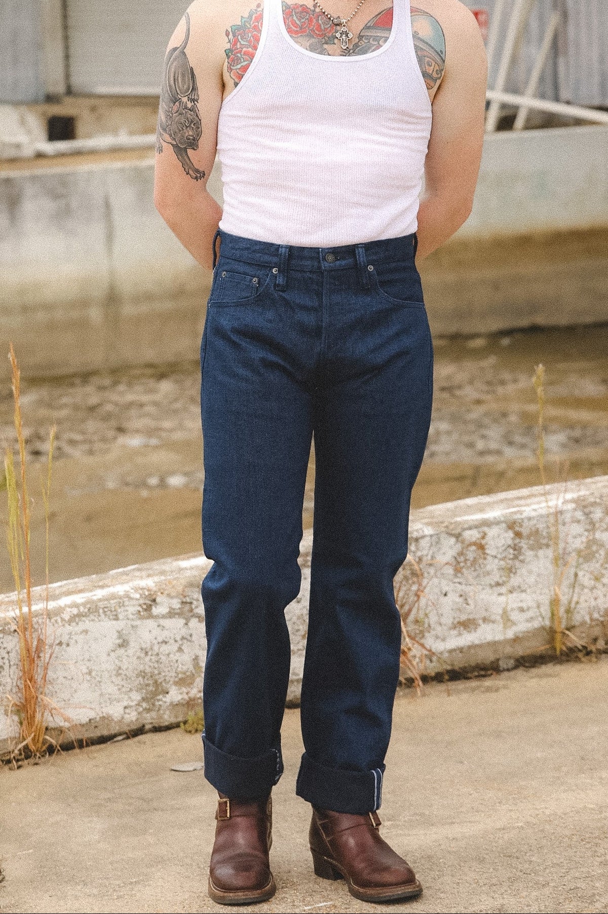 A man wearing dark blue straight-leg jeans, a white tank top, and brown leather boots, standing outdoors near a concrete canal.