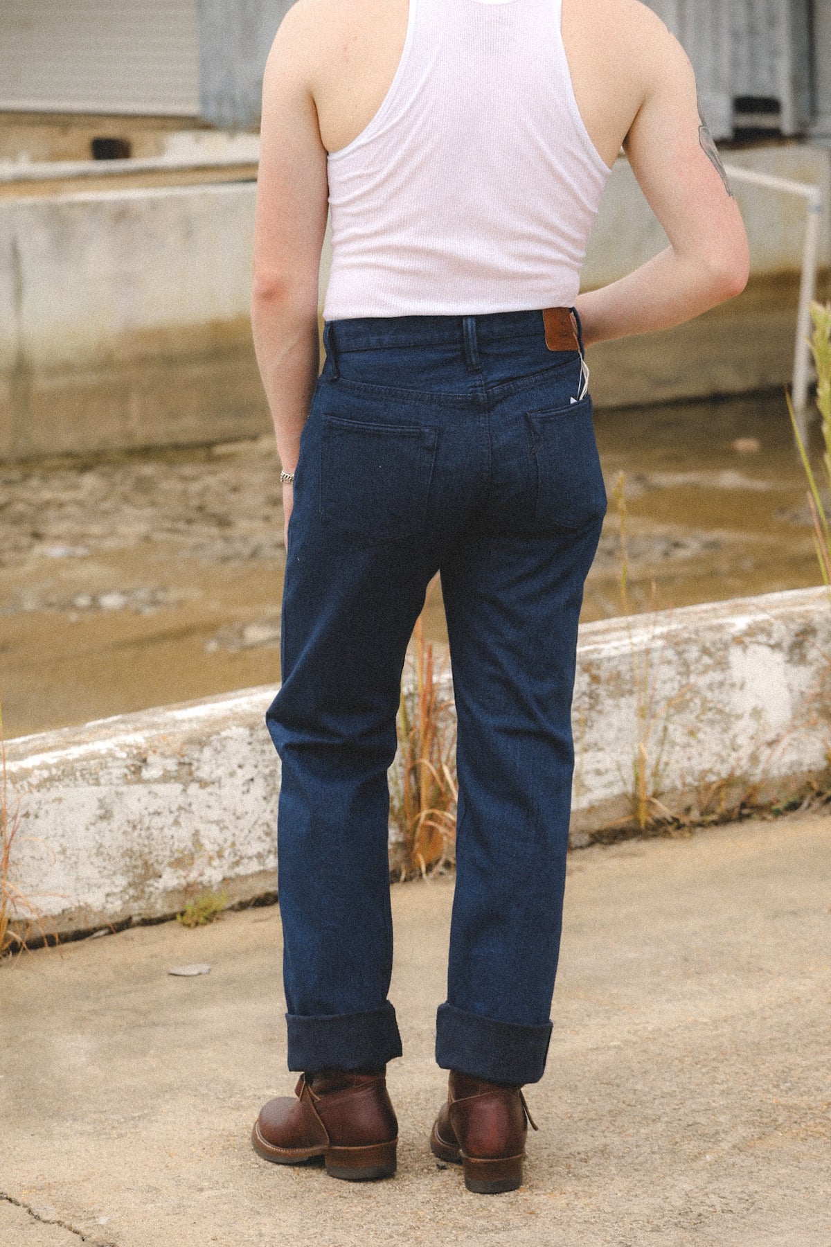 A person viewed from behind wearing dark blue denim jeans, a white tank top, and brown boots, standing outdoors on a concrete surface near a low wall.