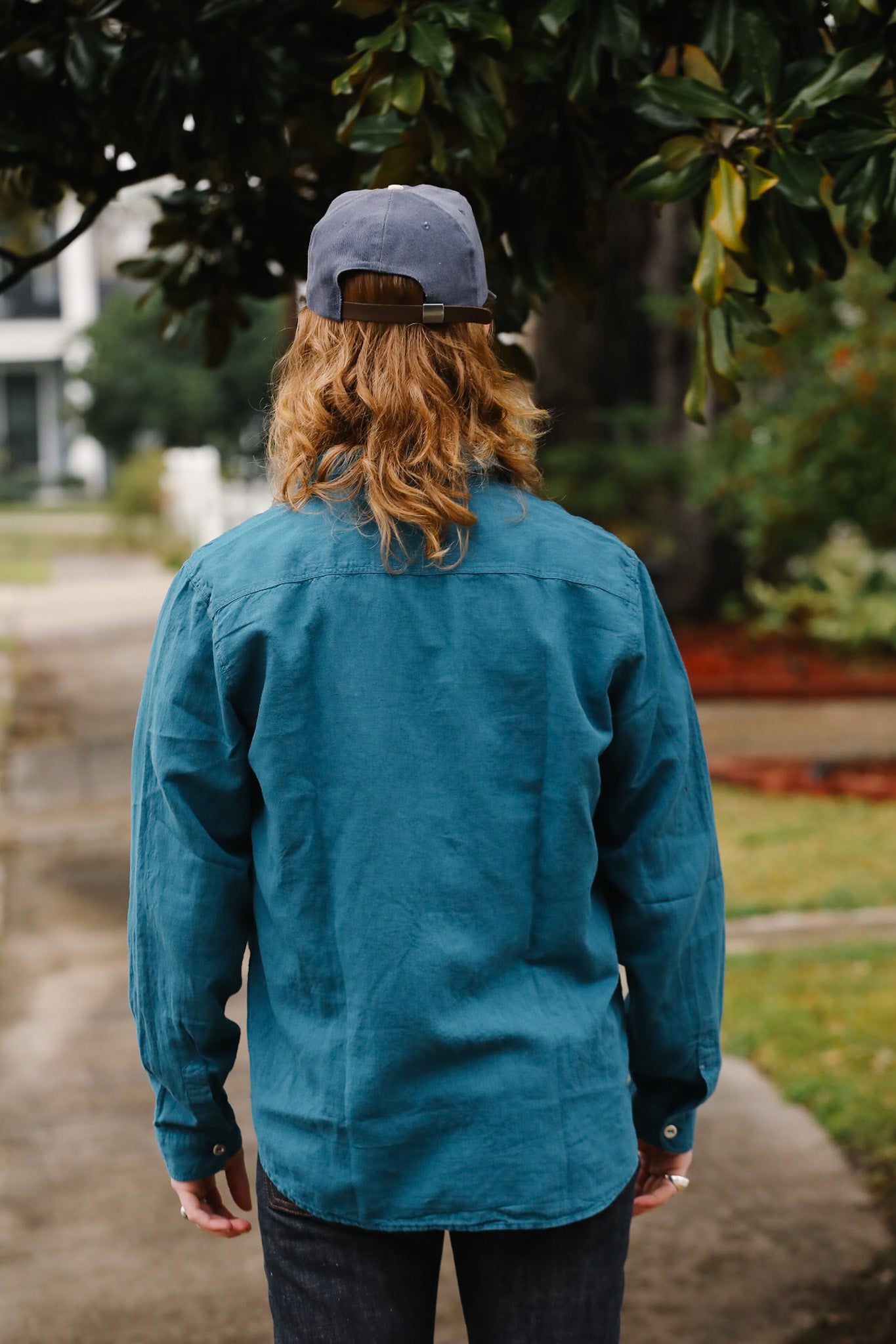 Person wearing a teal jacket and cap walking outdoors with trees and grass in the background