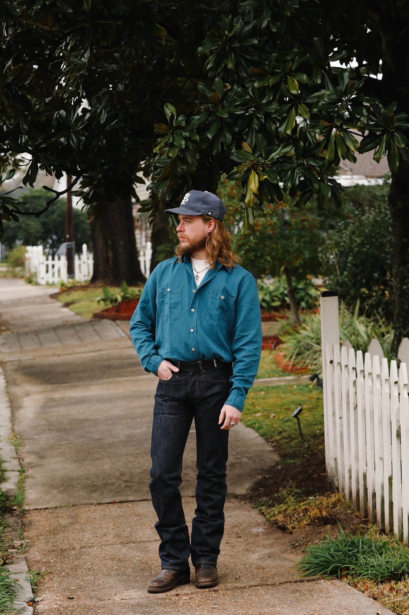 Man in a blue shirt and cap standing on a sidewalk with trees and a white picket fence in the background.