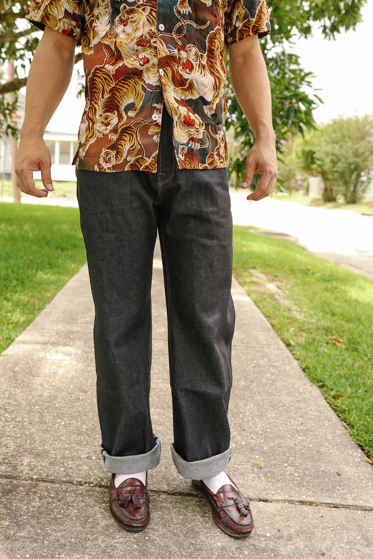 Man wearing dark raw denim jeans cuffed at the ankle with white socks and brown tassel loafers on a sidewalk
