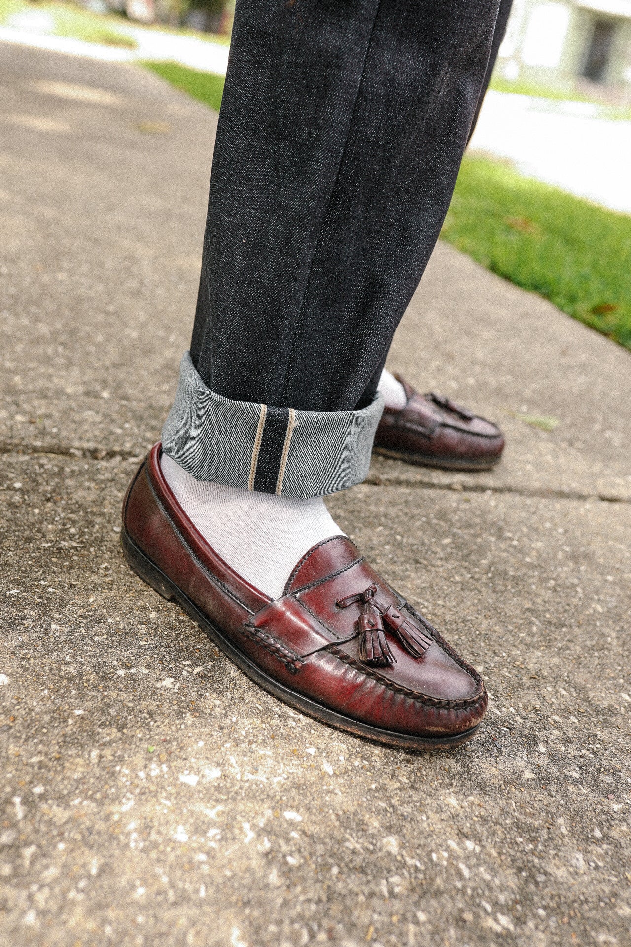 Close-up of rolled-up dark selvedge denim jeans paired with white socks and brown leather tassel loafers on sidewalk
