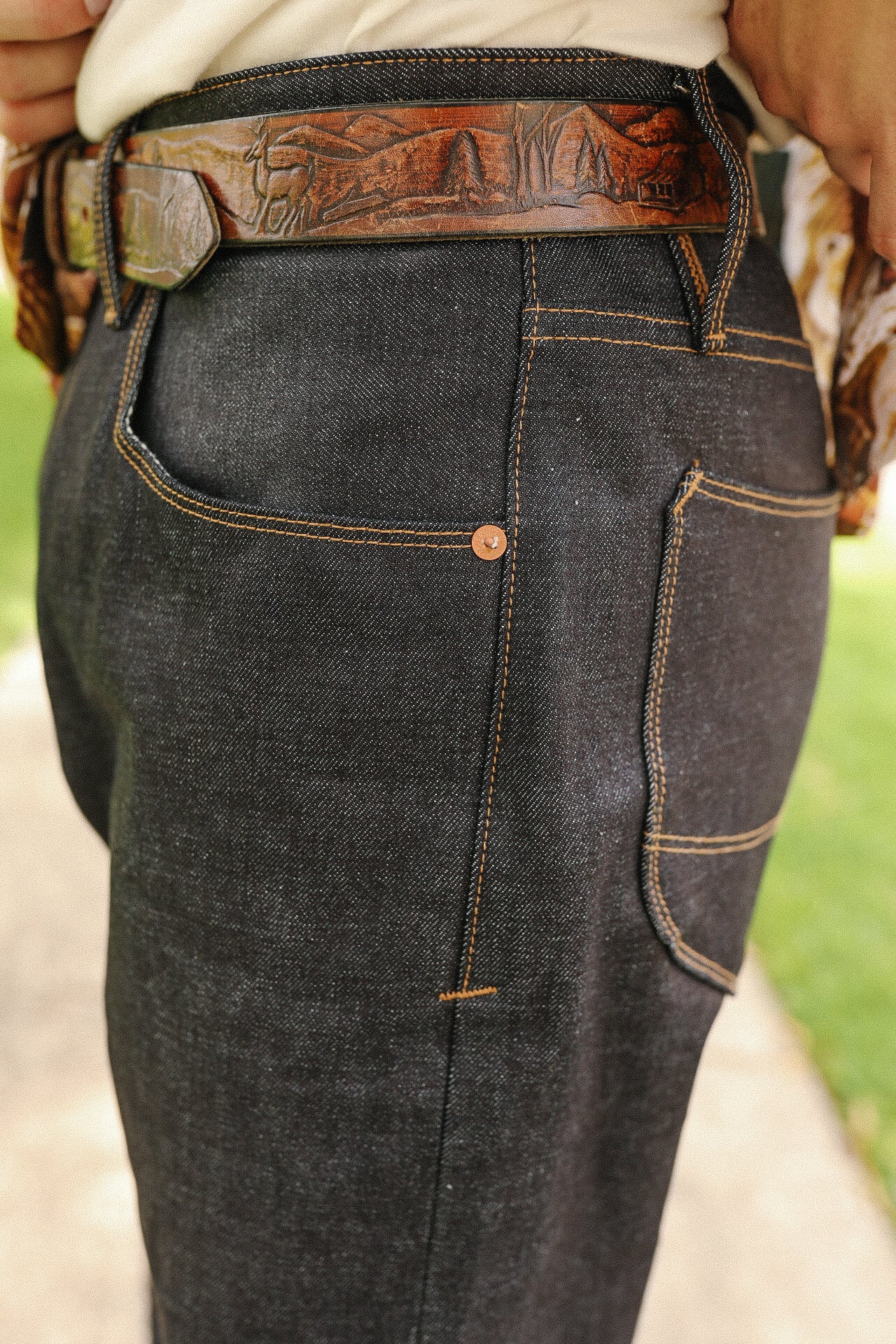 Close-up of dark raw denim jeans with contrast stitching and a brown leather belt featuring engraved mountain and deer design