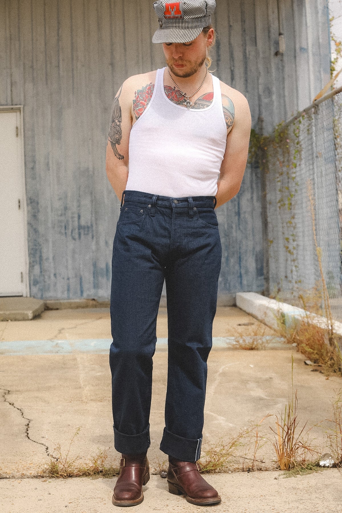 A man wearing a white tank top, dark blue denim jeans, and brown leather boots stands outdoors in front of a weathered gray metal wall. He is also wearing a gray baseball cap with a red logo.