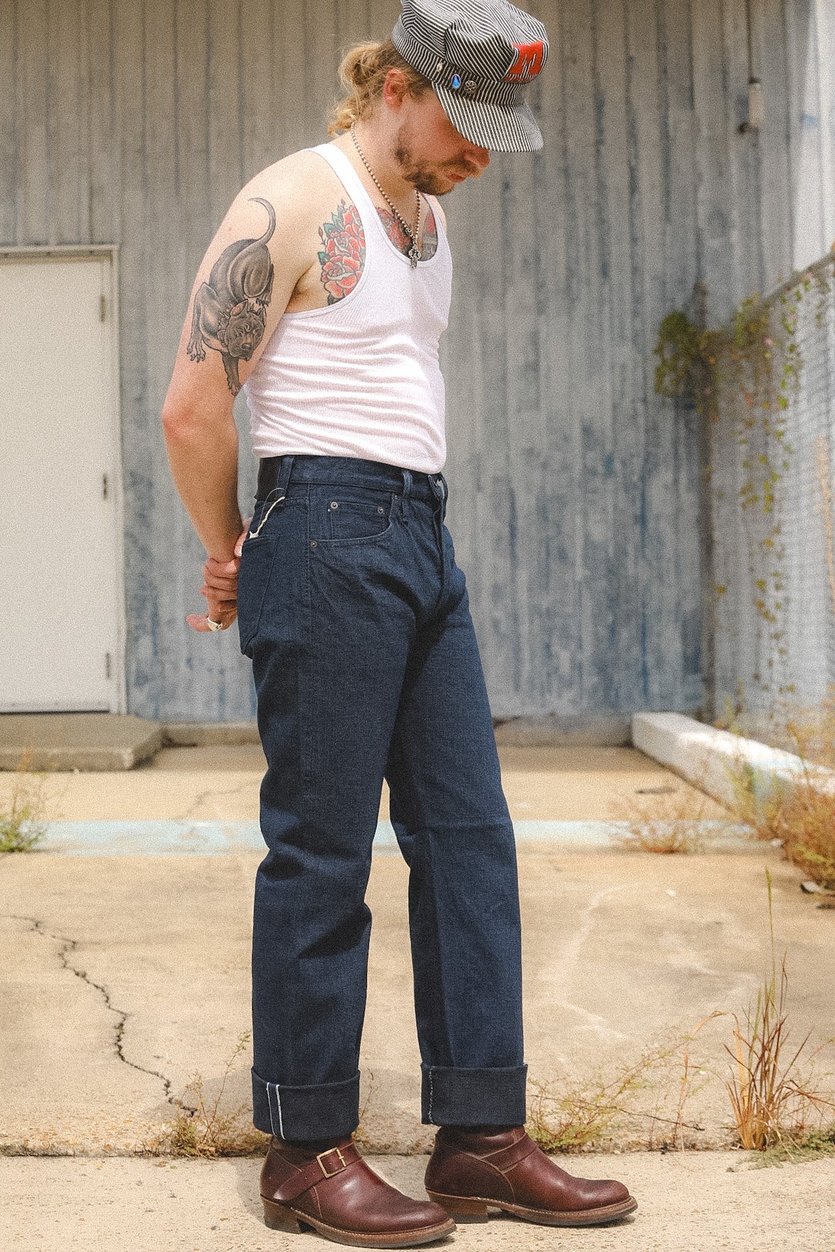 A man wearing dark blue denim jeans, a white tank top, brown leather boots, and a gray cap stands outdoors against a weathered gray metal wall.