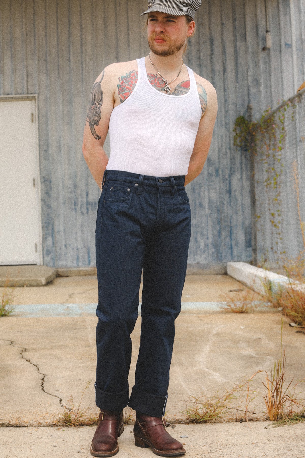 A man wearing a white ribbed tank top, dark blue jeans, and brown leather boots, standing outdoors in front of a weathered gray metal building.