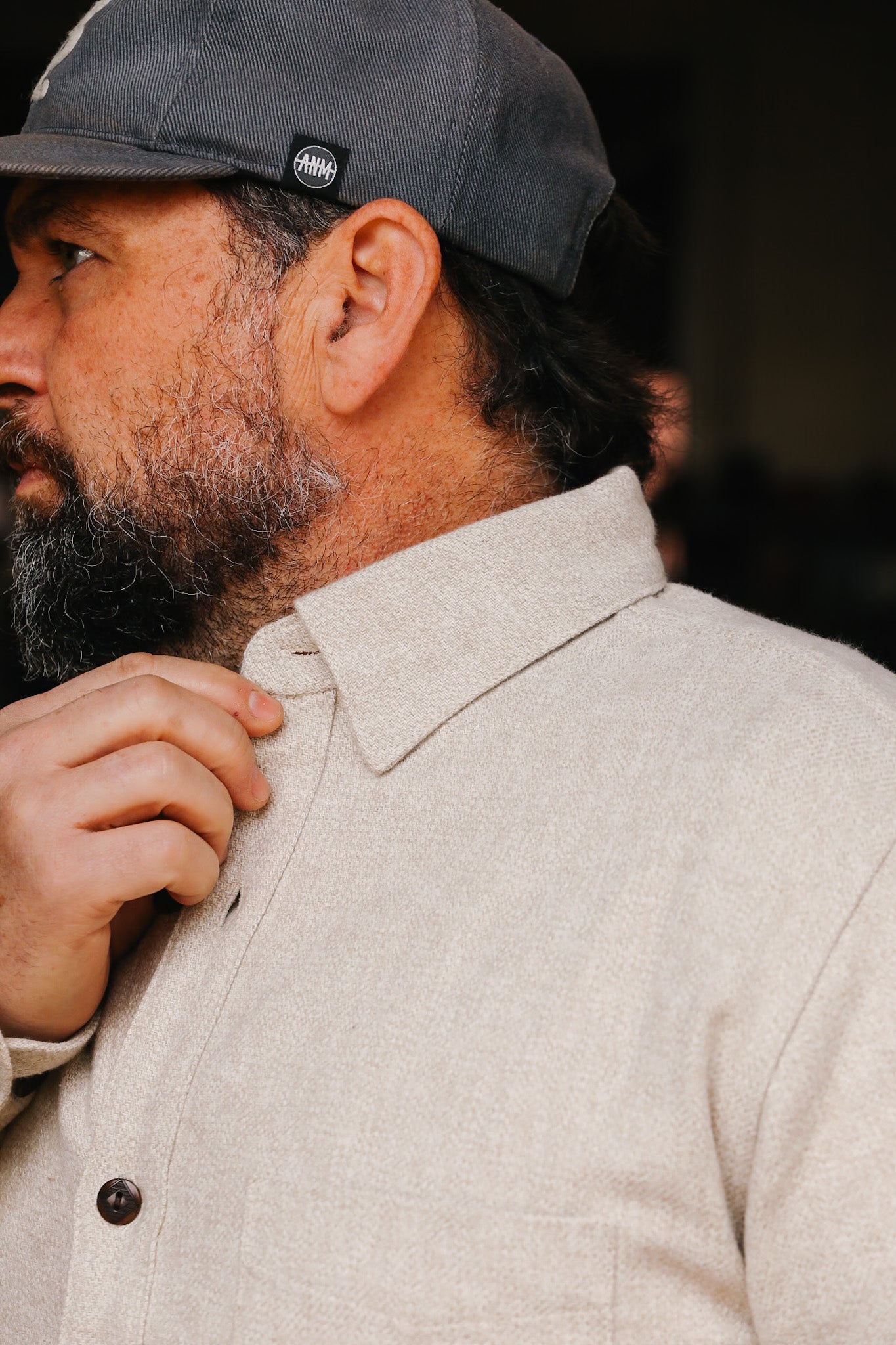 Bearded man adjusting collar of cream flannel Oxford shirt wearing dark cap