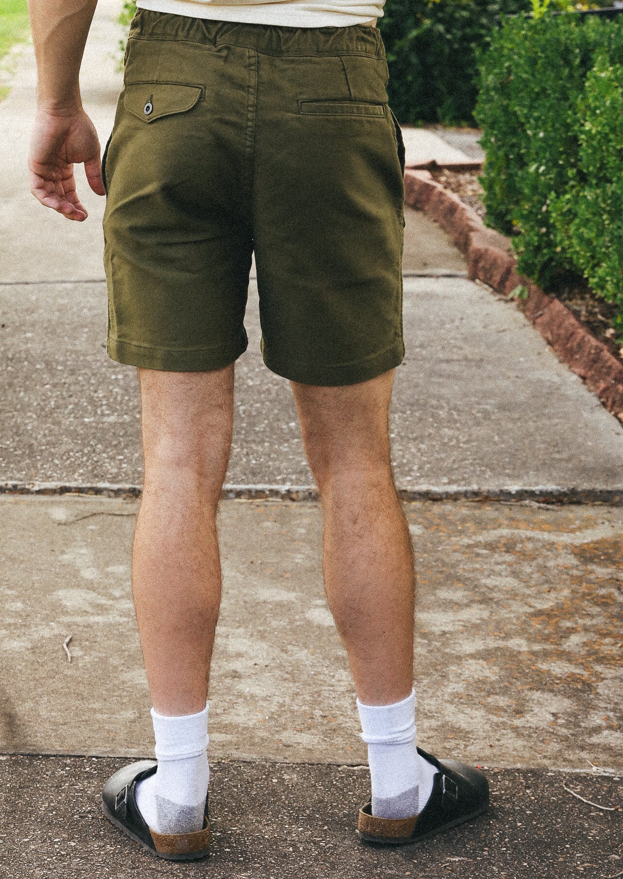 Man wearing olive green casual shorts, white socks, and black sandals standing on a sidewalk near greenery