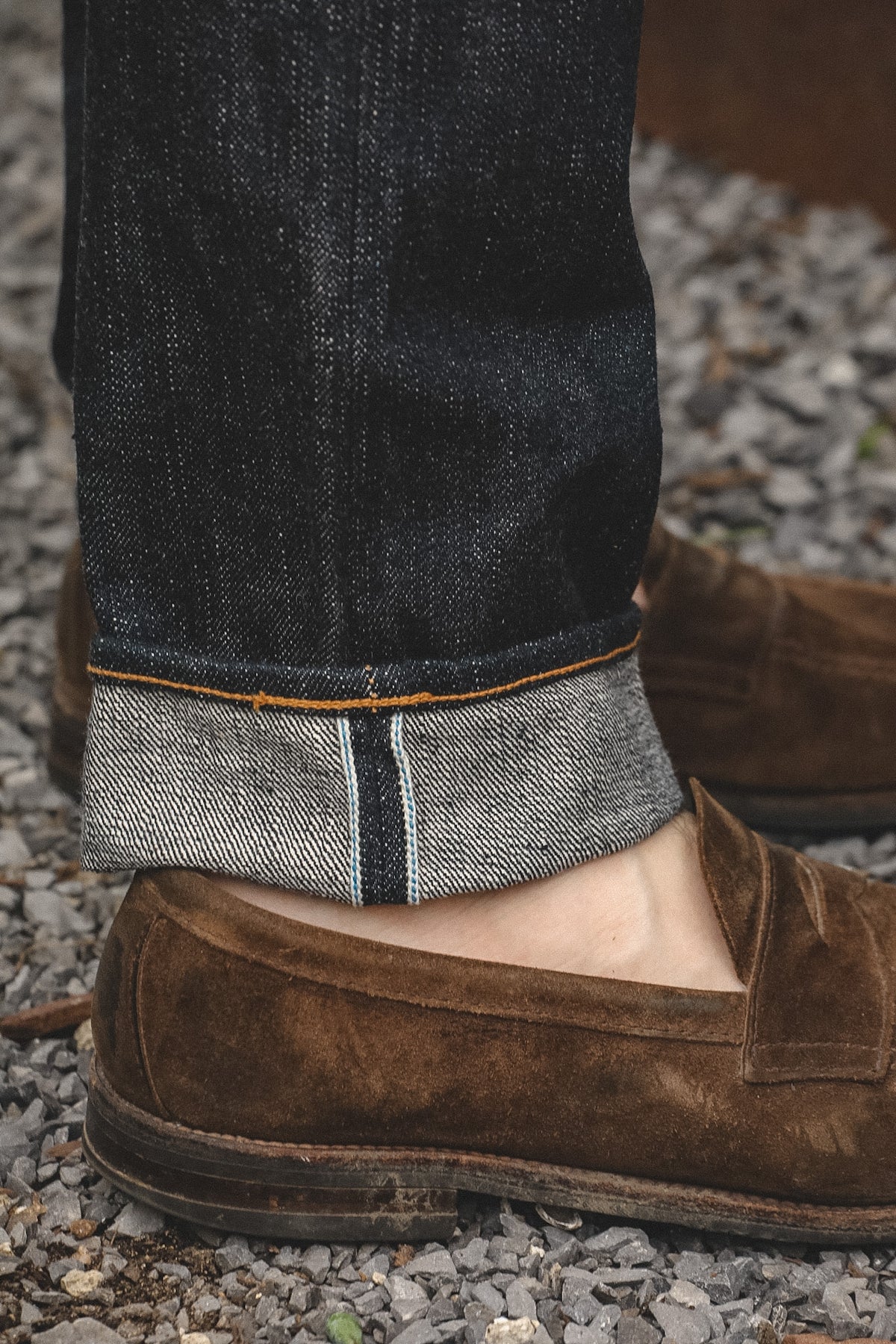 Close-up of a person's lower legs wearing dark indigo jeans with a turned-up hem showing tan and black contrast stitching, paired with brown suede penny loafers on a gravel surface.
