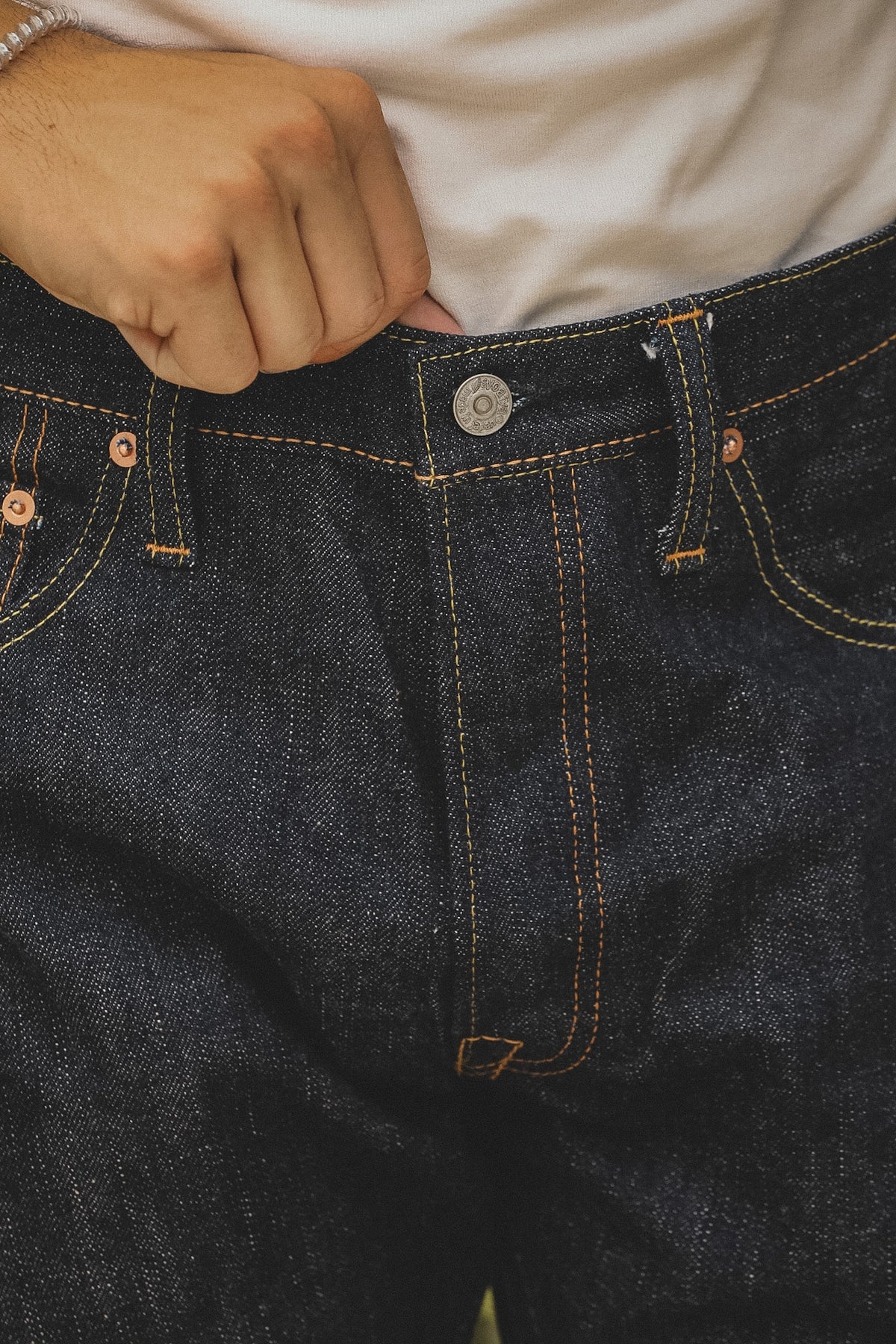 Close-up of a person wearing dark indigo denim jeans, with a hand pulling the waistband slightly upward over a white shirt.