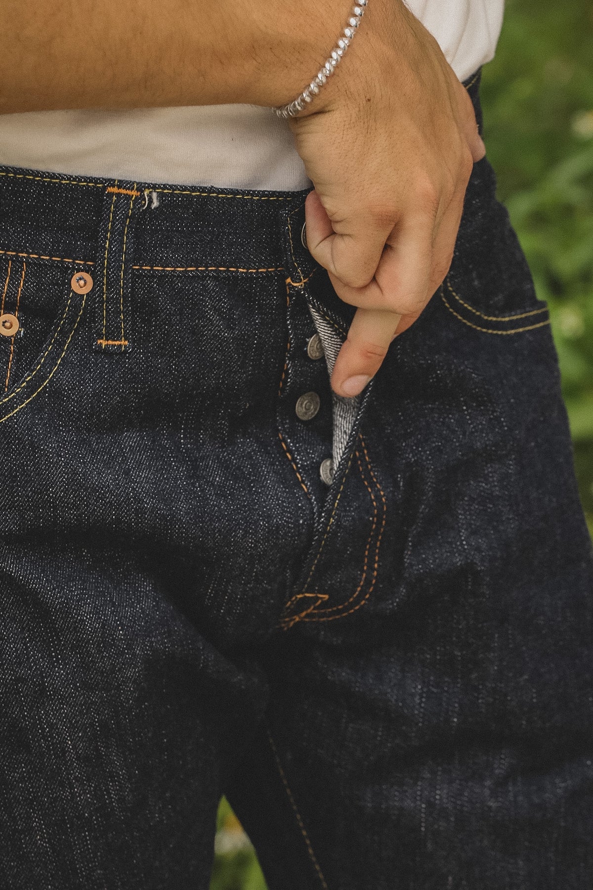 Close-up of a person wearing dark indigo denim jeans, pulling the waistband down slightly to reveal the inner lining and button area, with a white shirt and a silver bracelet visible.