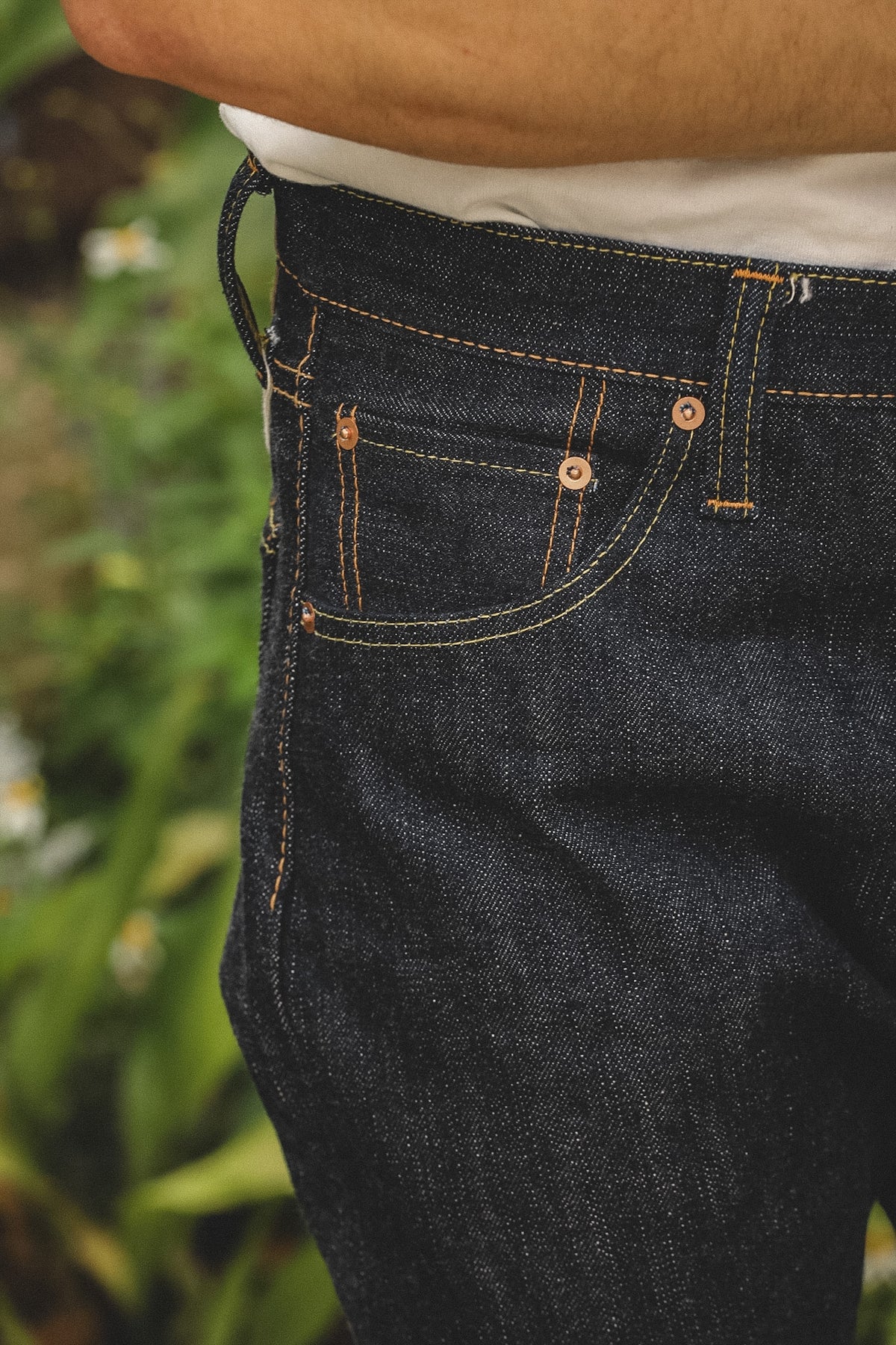 Close-up of a person wearing dark indigo denim jeans with tan contrast stitching, a white shirt, and copper-toned rivets, photographed outdoors with blurred green foliage in the background.