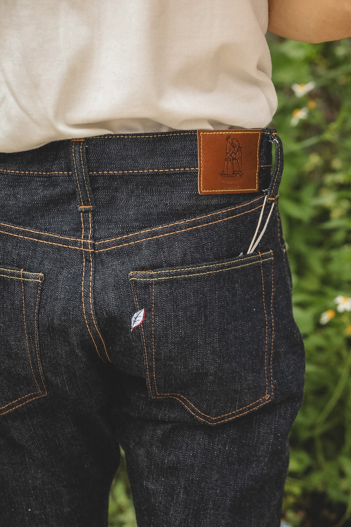 Close-up back view of a person wearing dark indigo denim jeans with contrast stitching, a brown leather patch at the waistband, and a small white embroidered logo on the back pocket, set against a blurred green outdoor background.