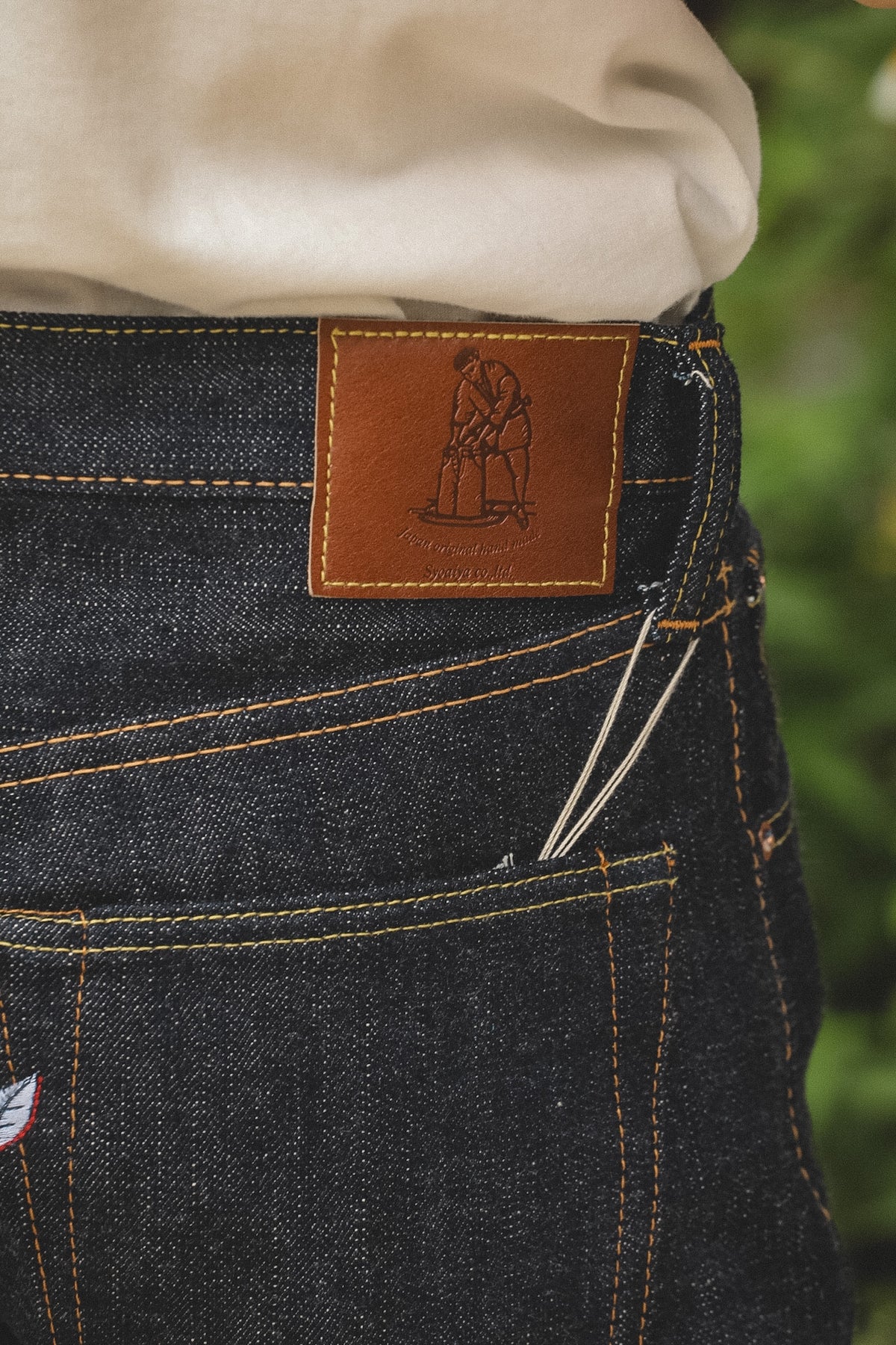Close-up of the back waistband of dark indigo denim jeans with a brown leather patch showing the 'Hagasa' brand logo, worn with a cream-colored shirt.