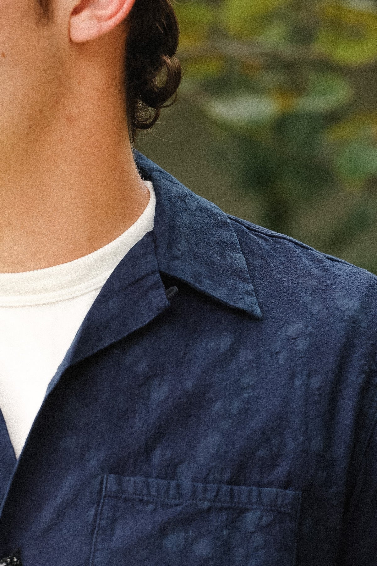 Close-up of a person wearing a textured navy blue overshirt over a white t-shirt, with a blurred outdoor background.