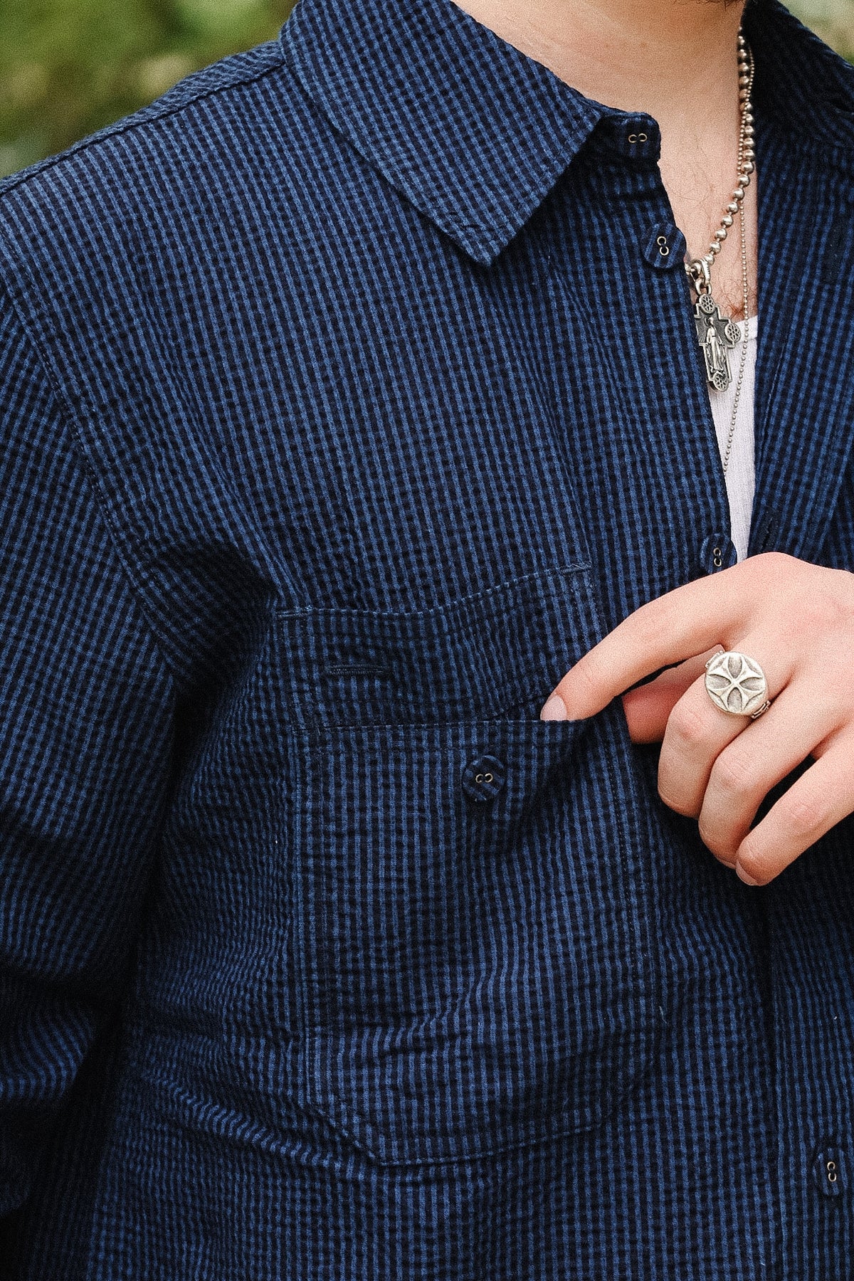 Close-up of a person wearing a dark navy blue and black micro-check patterned button-up shirt, with a silver chain necklace and a silver ring with a white stone on the hand.