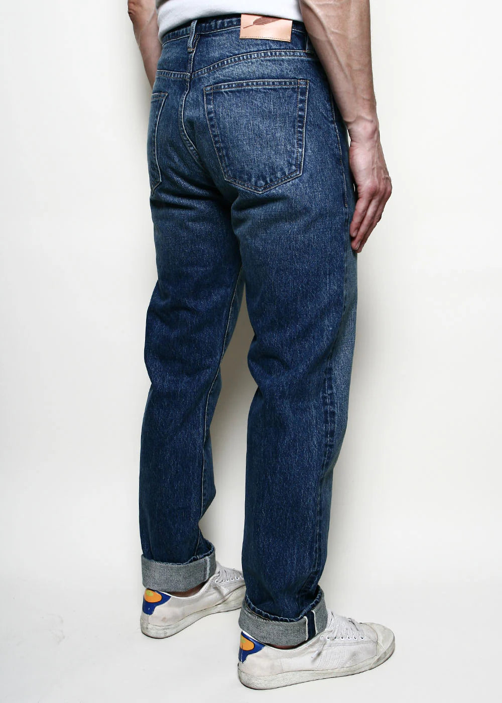 Back view of a person wearing dark blue rolled-up denim jeans and white sneakers against a plain white background