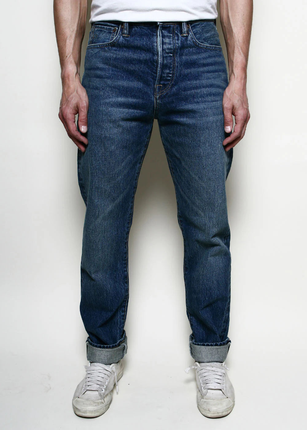 Front view of a person wearing dark blue rolled-up jeans and white sneakers against a plain background