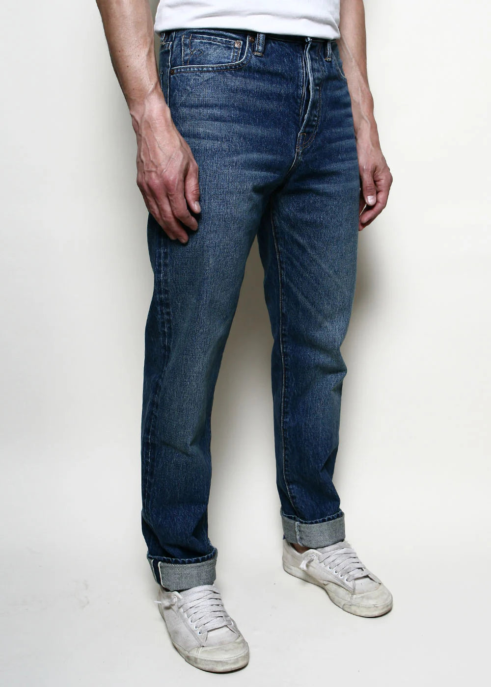 Man wearing rolled-up dark blue denim jeans and white sneakers against a plain background