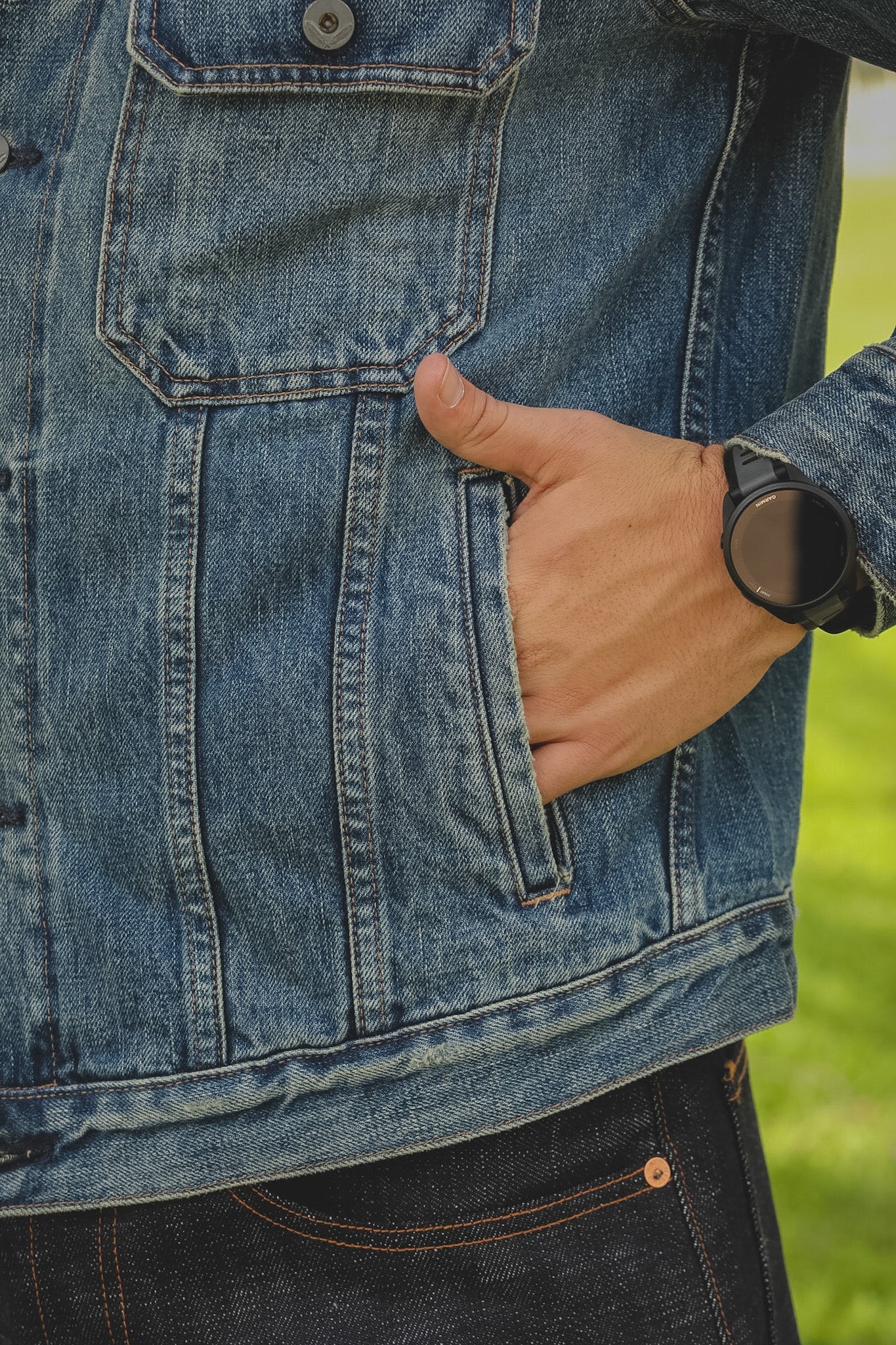 Person wearing a blue denim jacket with a hand in the pocket, standing against a green outdoor background.