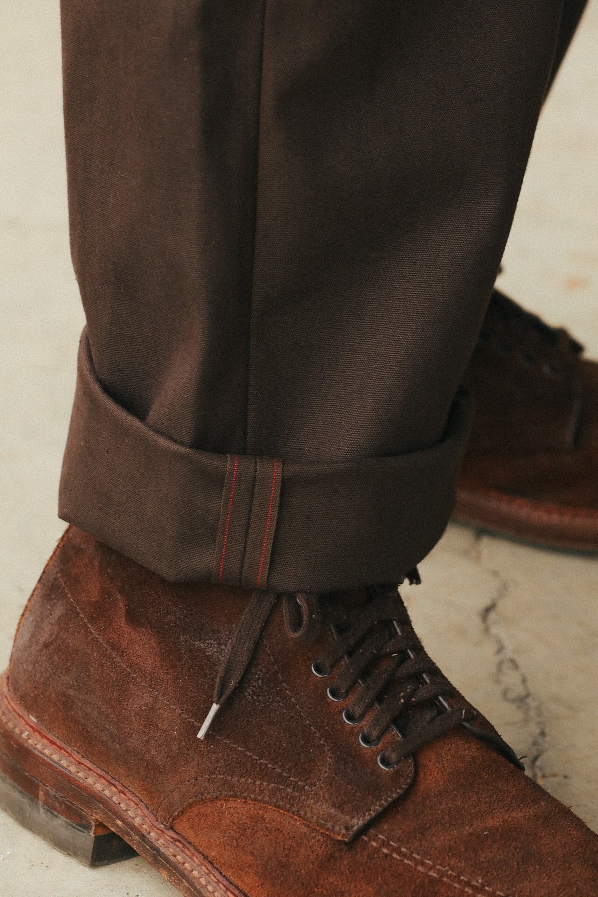 Close-up of dark brown trousers with a rolled cuff and dark brown suede lace-up shoes on a light stone floor.