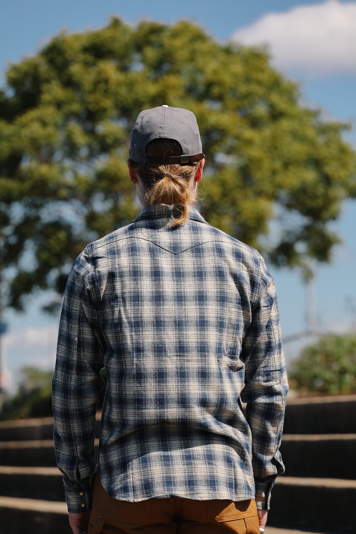 A man viewed from behind wearing a blue and white plaid flannel shirt, a gray baseball cap, and brown pants, standing outdoors in front of a large green tree and blue sky.