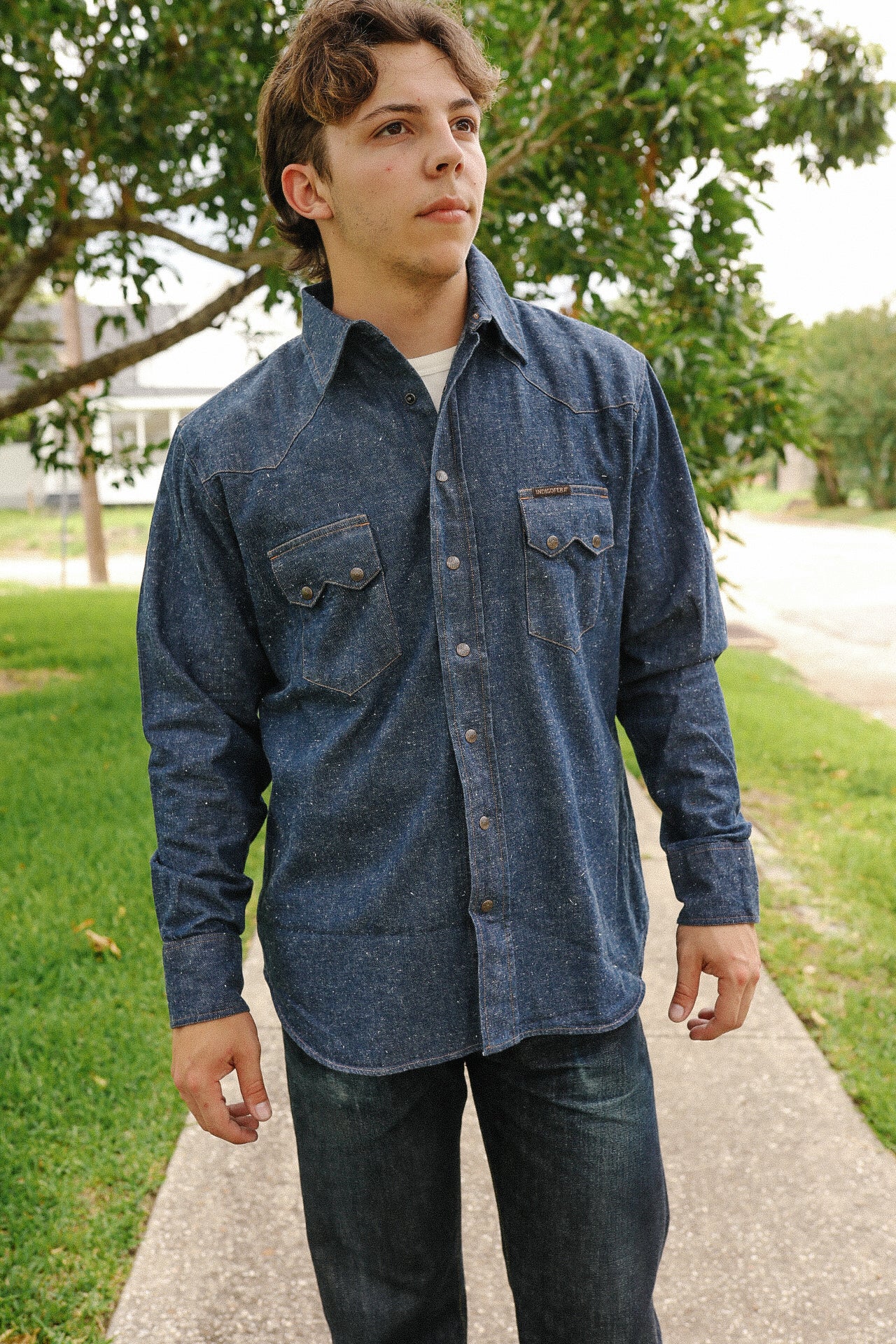 Young man wearing neppy indigo denim shirt with buttoned chest pockets outdoors by green grass and trees