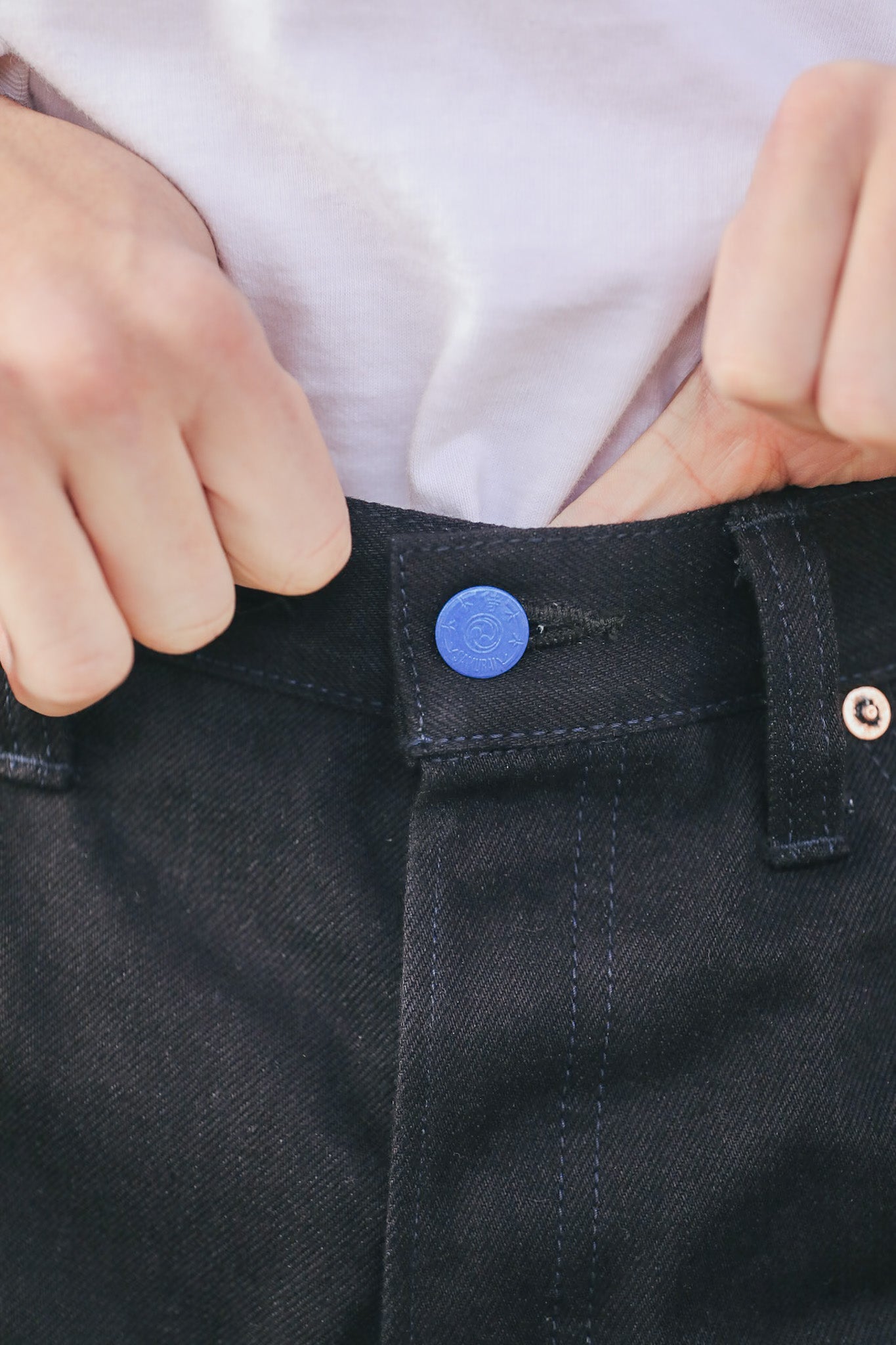 Close-up of a person adjusting a button on dark jeans with a white shirt background.